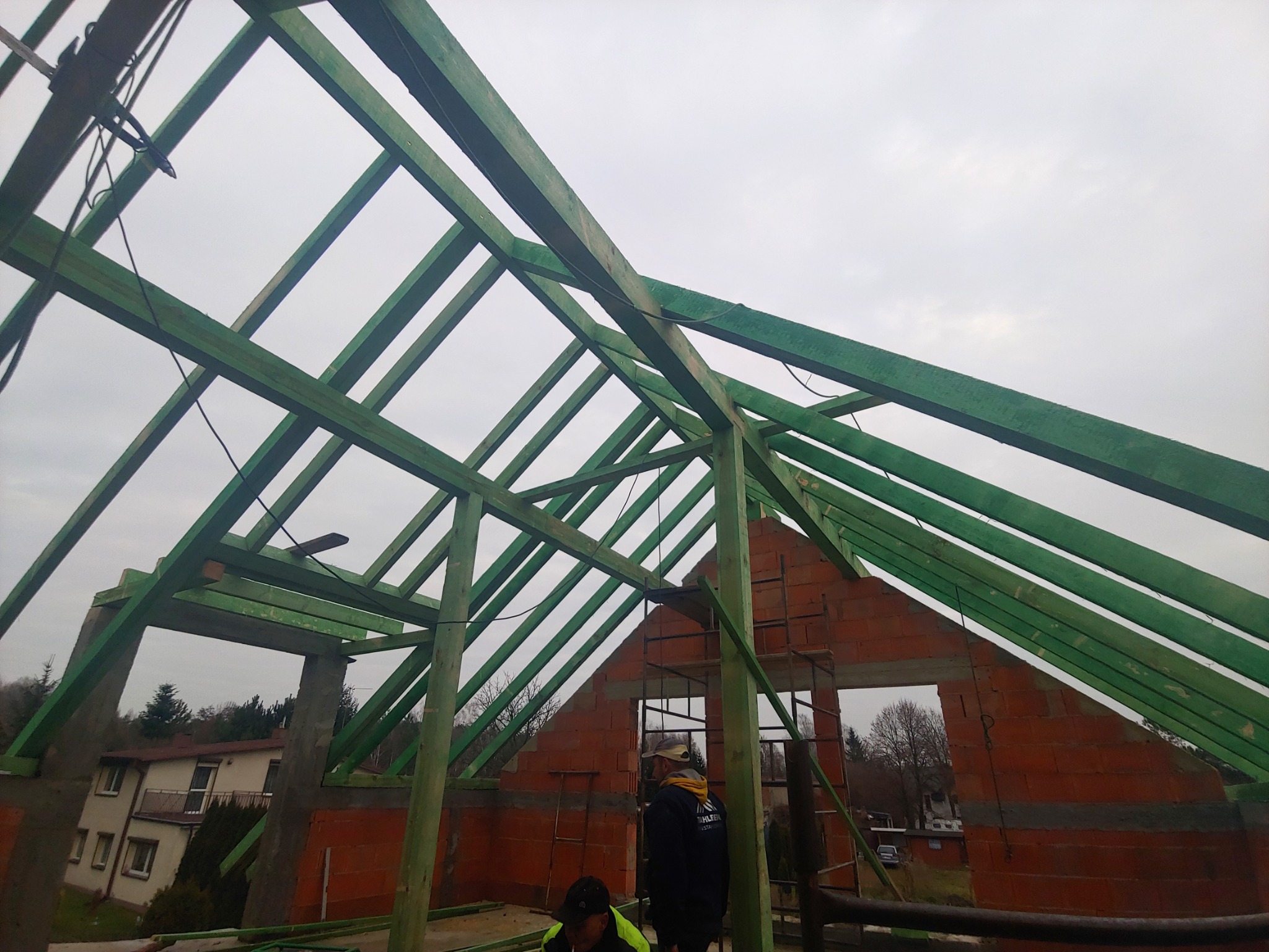 Konstrukcja drewniana więźby dachowej z zielonych belek na murowanym budynku w trakcie budowy, widoczne dwie osoby w ubraniach roboczych.