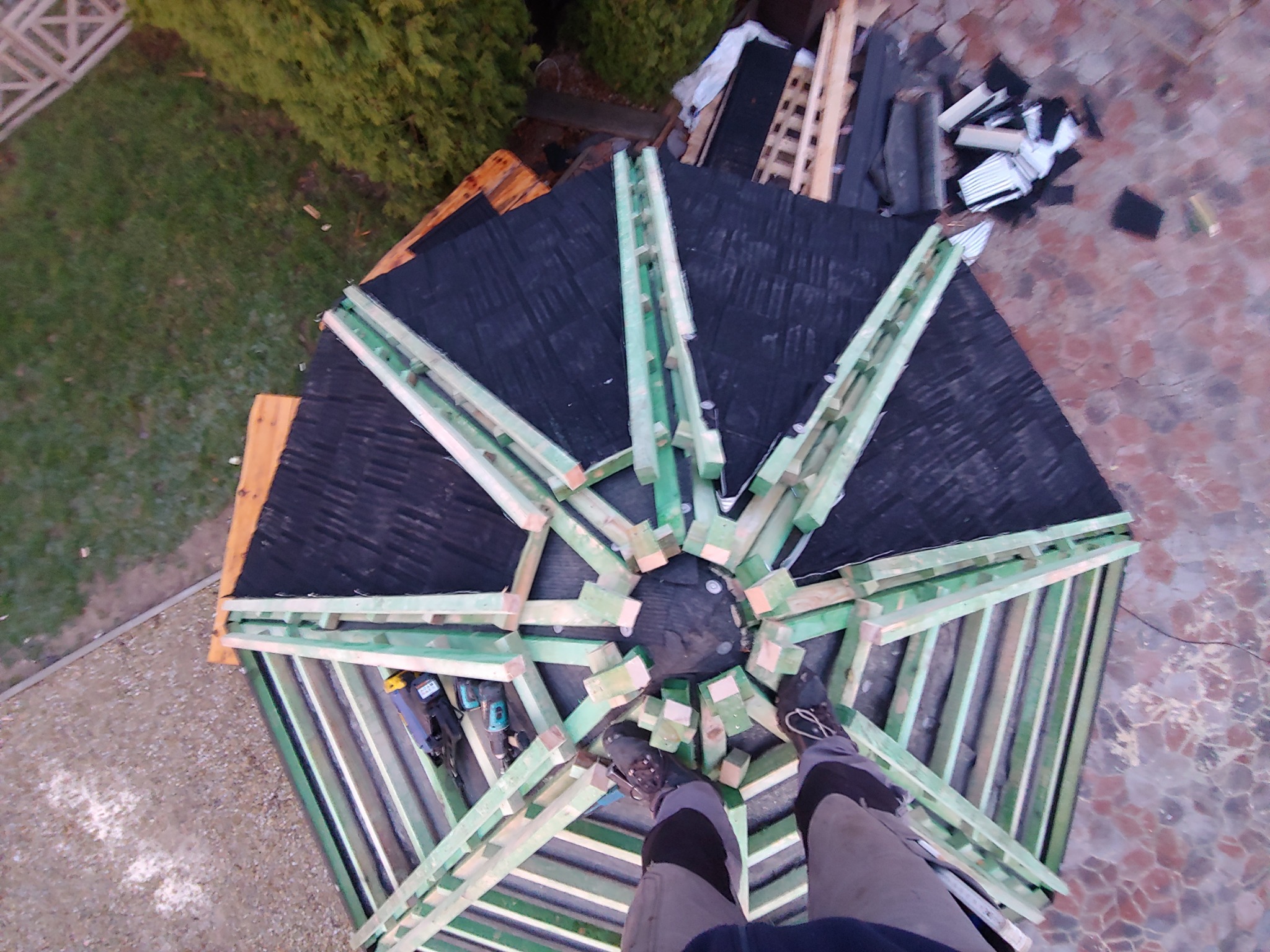 Widok z góry na konstrukcję dachu altany w trakcie budowy, z widocznymi elementami drewnianymi, czarną papą i narzędziami, oraz stopami dekarza w butach roboczych.