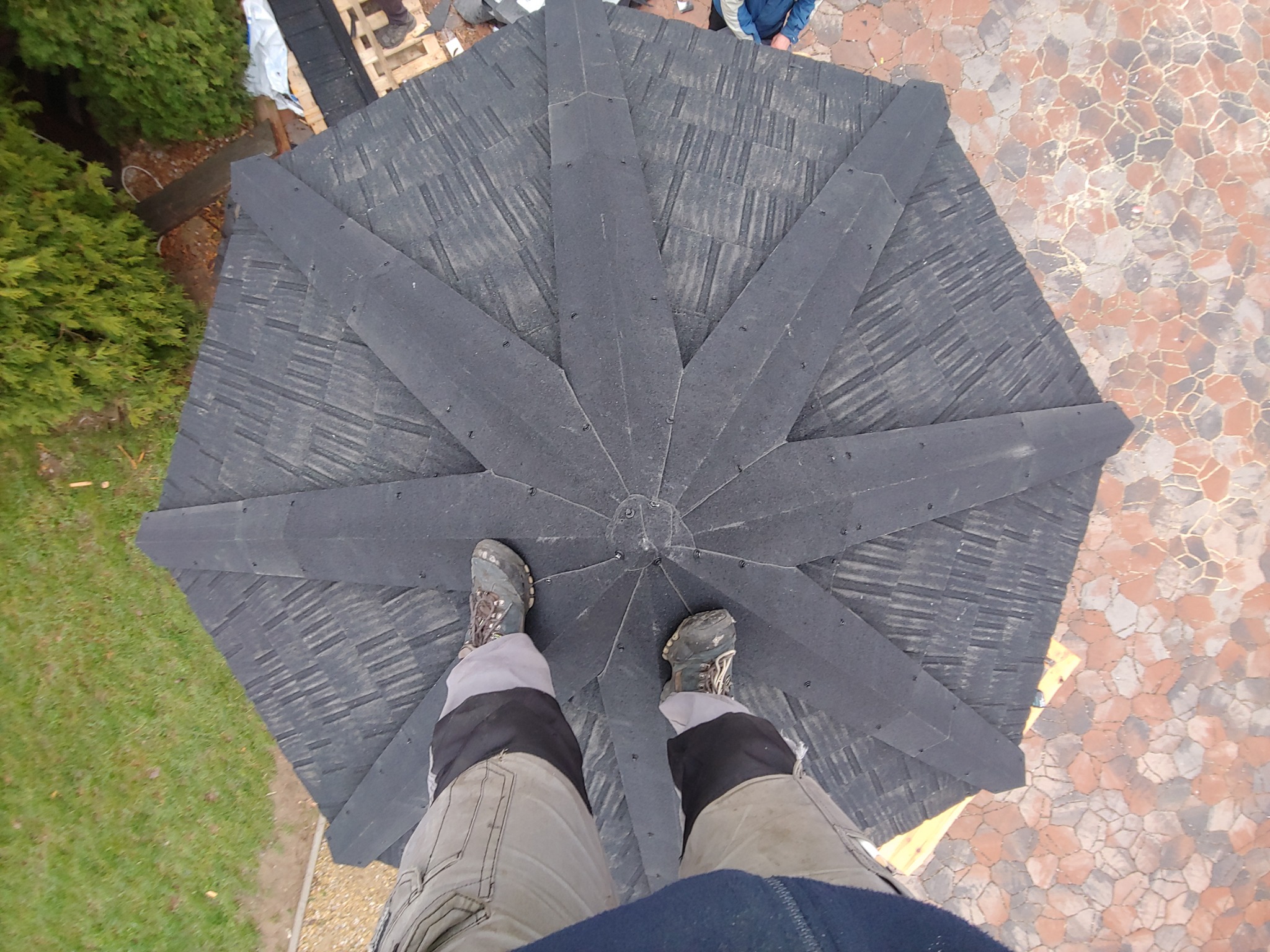 Widok z góry na ośmiokątny dach altany pokryty czarnym gontem bitumicznym, z widocznymi stopami dekarza w roboczych butach i spodniach, otoczenie z zielenią i kostką brukową.