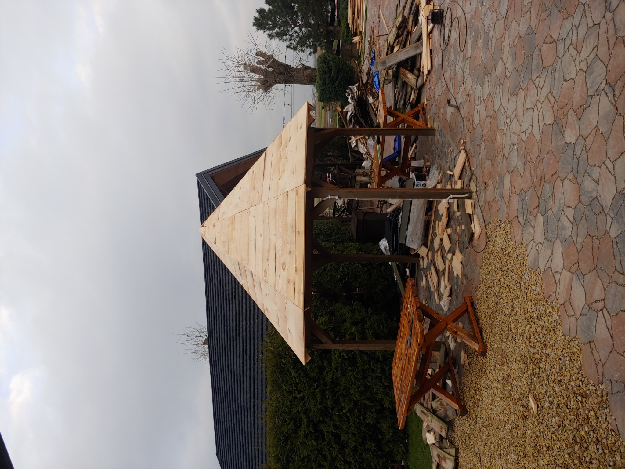 Konstrukcja drewnianej altany w trakcie budowy, widoczne deski tworzące zadaszenie, fragment dachu domu z czarną blachodachówką, porozrzucane drewniane elementy i narzędzia na brukowanej powierzchni.