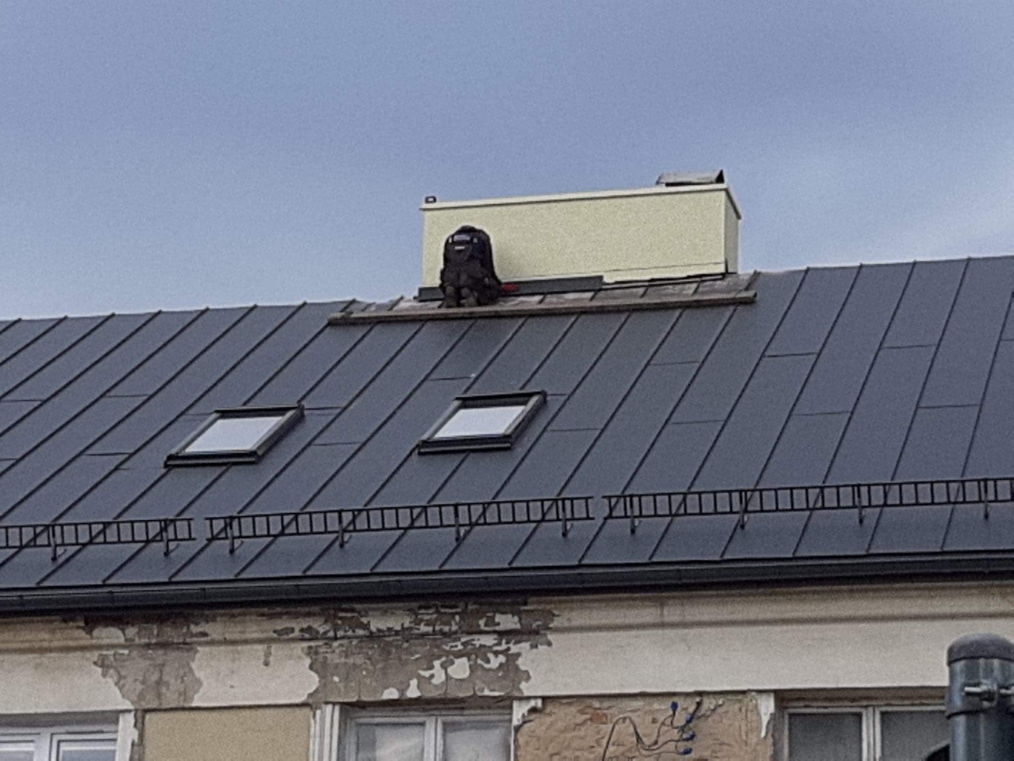 Dekarz w ciemnym kombinezonie pracuje na dachu z blachy w kolorze grafitowym, widoczne dwa okna dachowe i komin w jasnym kolorze