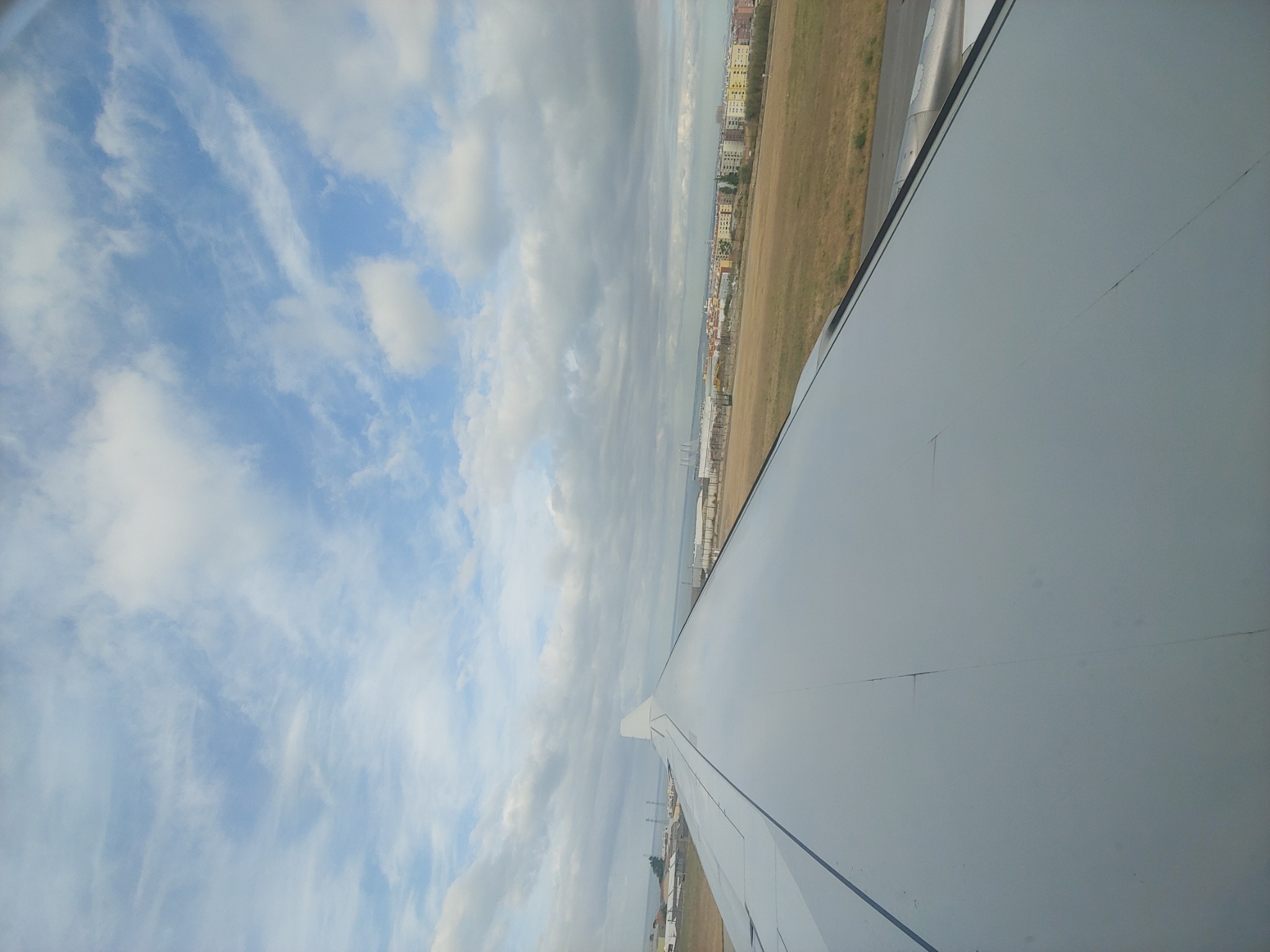 Widok z okna samolotu na skrzydło, chmury i zabudowania miejskie w oddali.