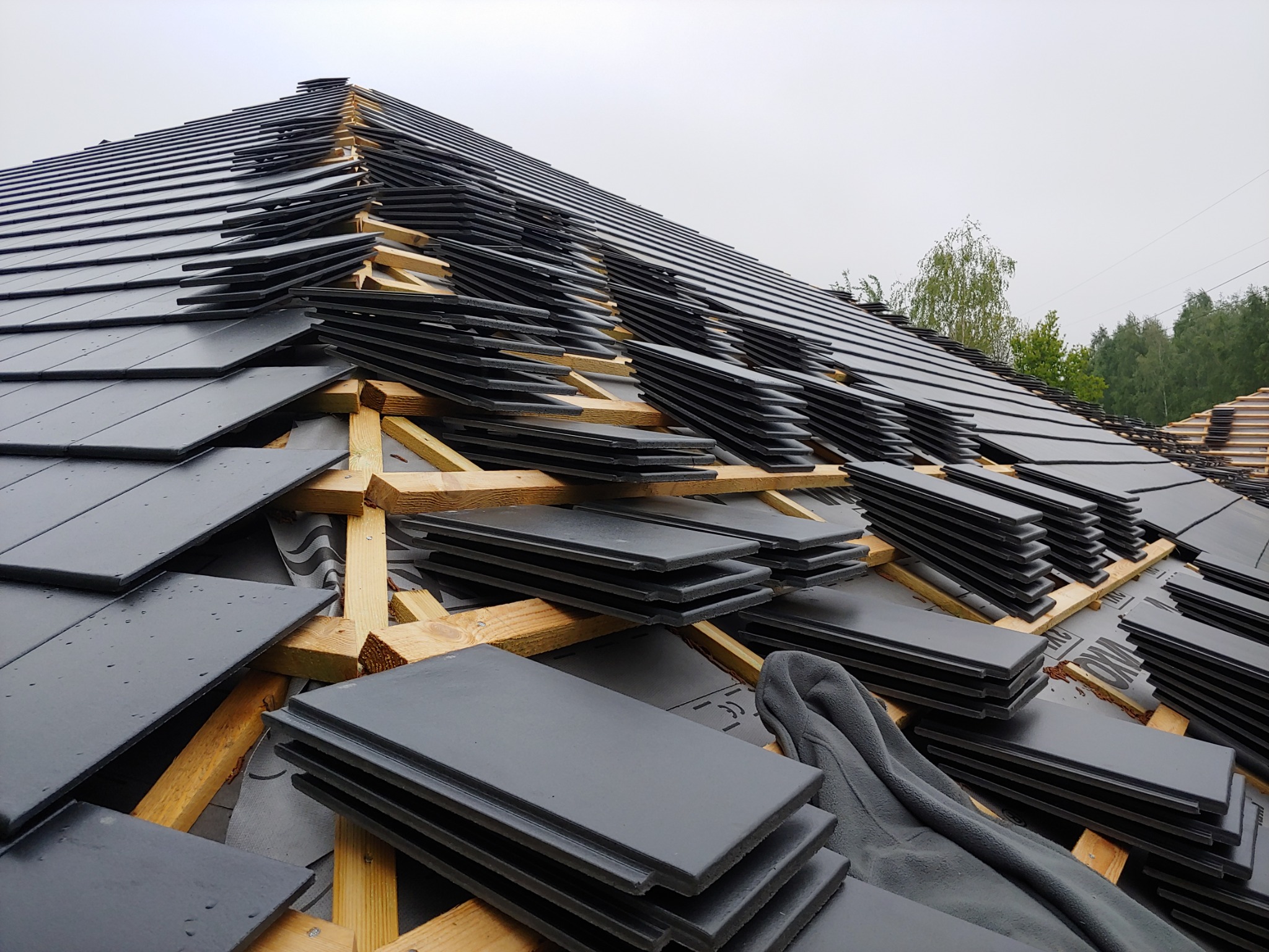 Układanie czarnej dachówki ceramicznej na drewnianej konstrukcji dachu, widoczne membrany dachowe i stosy płytek, pochmurne niebo w tle.