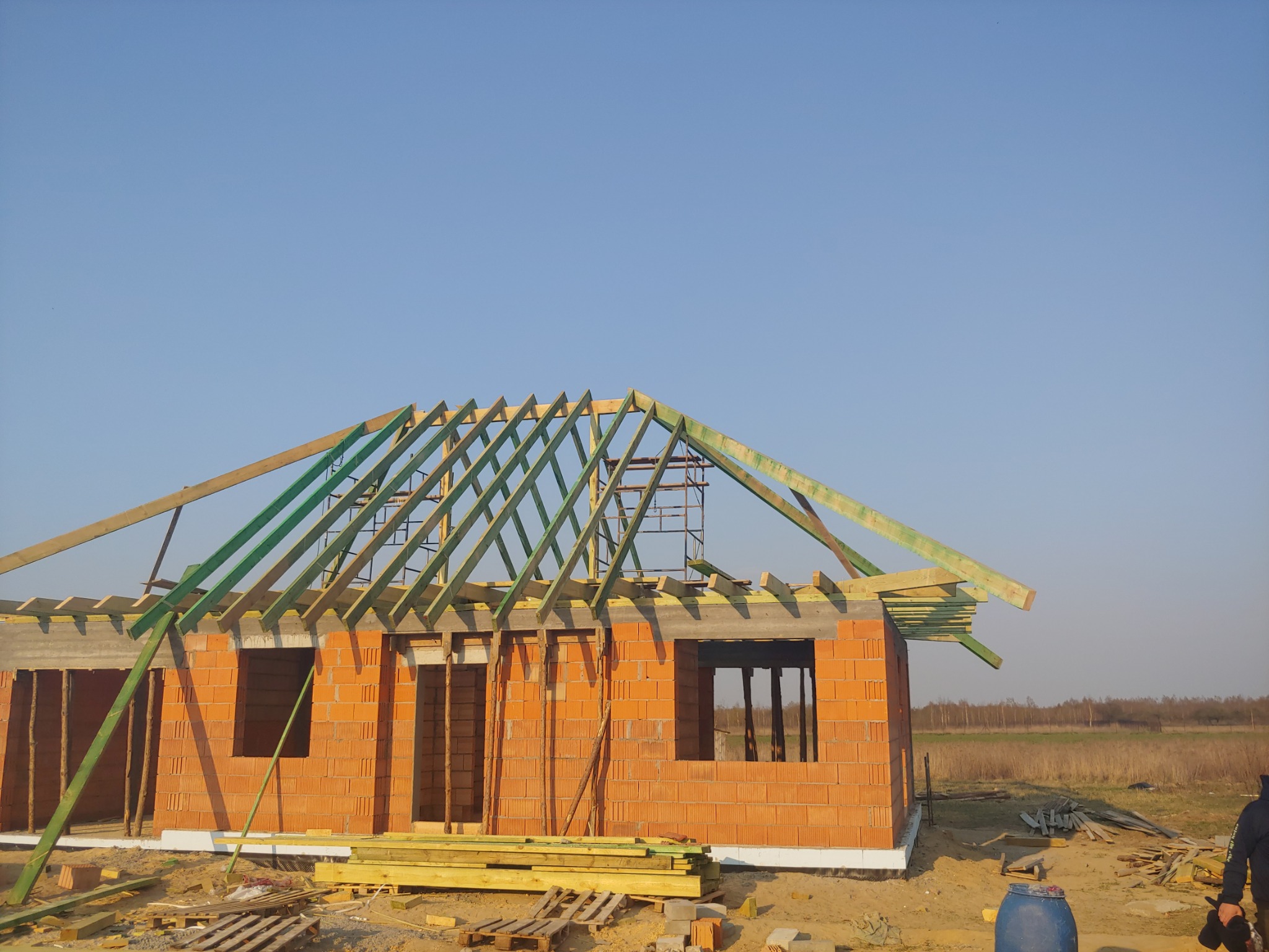 Surowa konstrukcja domu z czerwonej cegły z widoczną drewnianą więźbą dachową pomalowaną na zielono, w tle błękitne niebo i jesienny krajobraz.