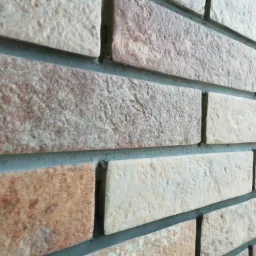 Ściana wyłożona dekoracyjną cegłą w odcieniach beżu i brązu, z widoczną szarą fugą, ułożona w regularny wzór, ujęcie pod kątem.