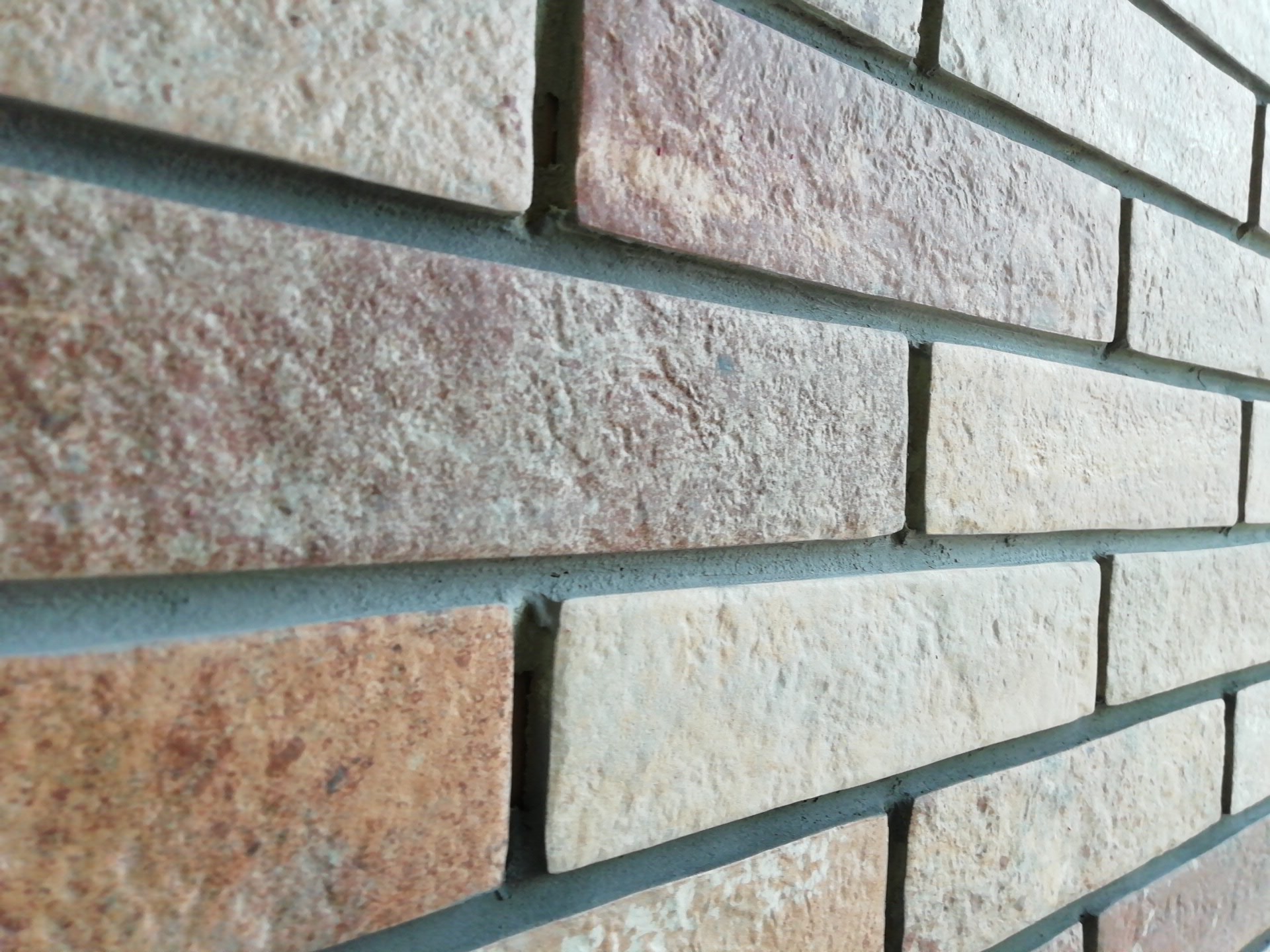 Ściana wyłożona dekoracyjną cegłą w odcieniach beżu i brązu, z widoczną szarą fugą, ułożona w regularny wzór, ujęcie pod kątem.