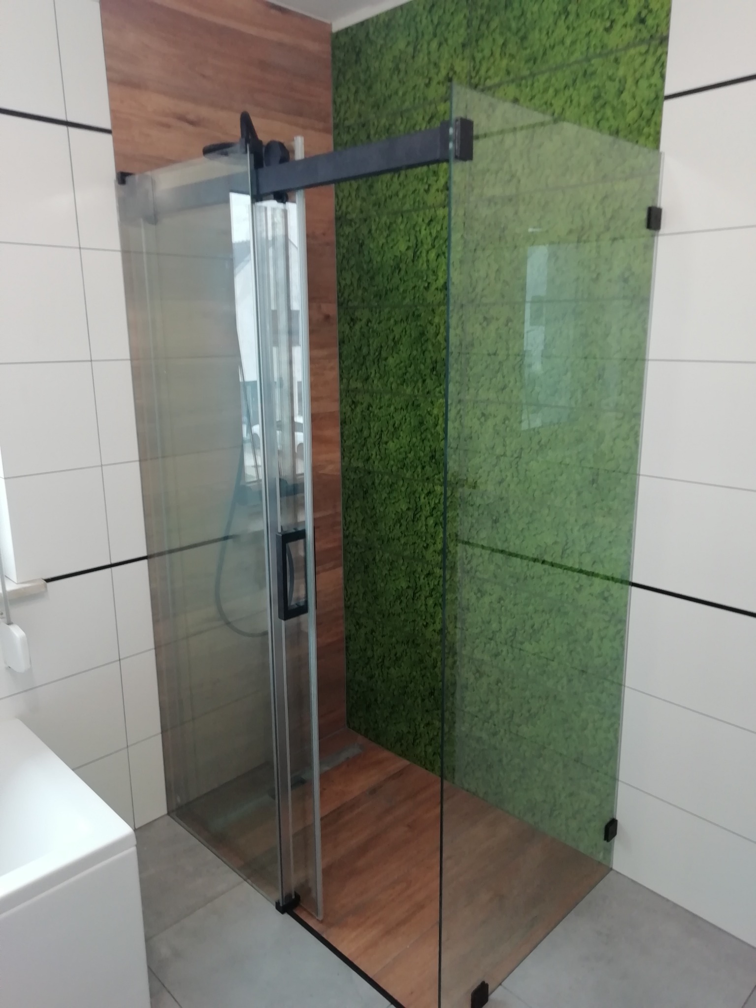 Nowoczesna kabina prysznicowa z przesuwnymi drzwiami, czarne profile, drewno i zielona ściana z mchu w tle, białe płytki na ścianach.