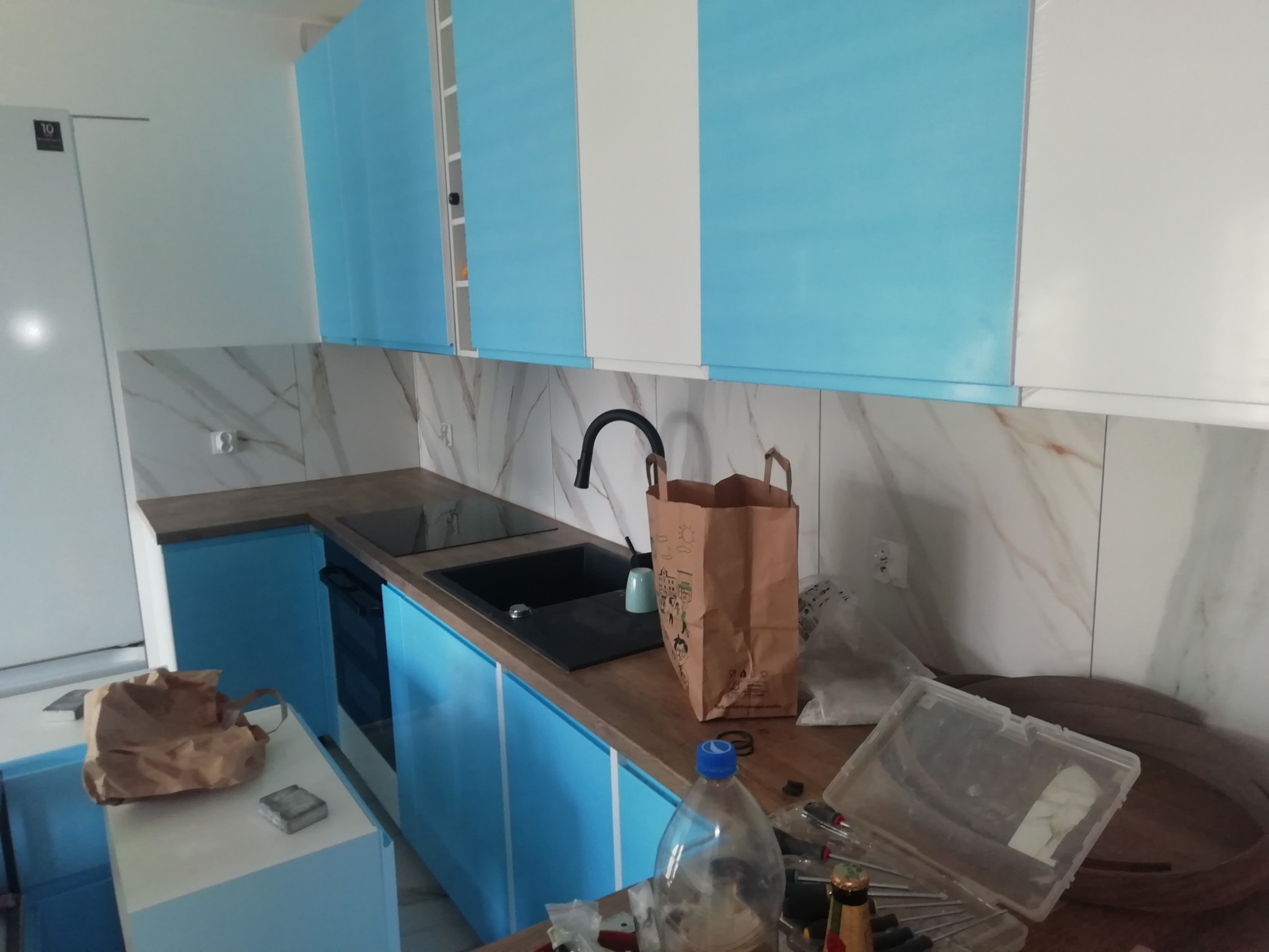 Kuchnia w trakcie montażu: błękitno-białe szafki wiszące i dolne, czarny zlew i płyta indukcyjna w blacie, marmurowe płytki na ścianie, widoczne narzędzia i akcesoria montażowe.