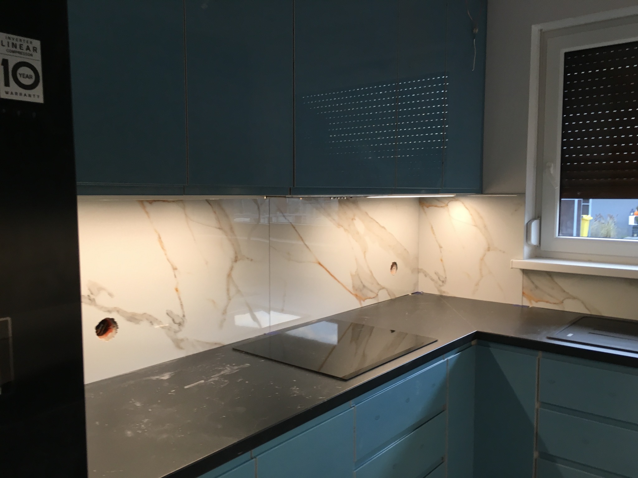 Fragment kuchni w trakcie montażu: błękitne szafki, czarny blat, płyta indukcyjna i białe panele ścienne z marmurowym wzorem, widoczne otwory na gniazdka.