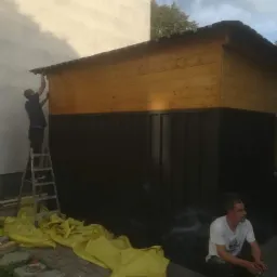 Pracownik na drabinie montuje metalowy element przy drewnianej konstrukcji, dół konstrukcji pomalowany na ciemny kolor, obok siedzi mężczyzna z papierosem, podłoże wyłożone kostką brukową.