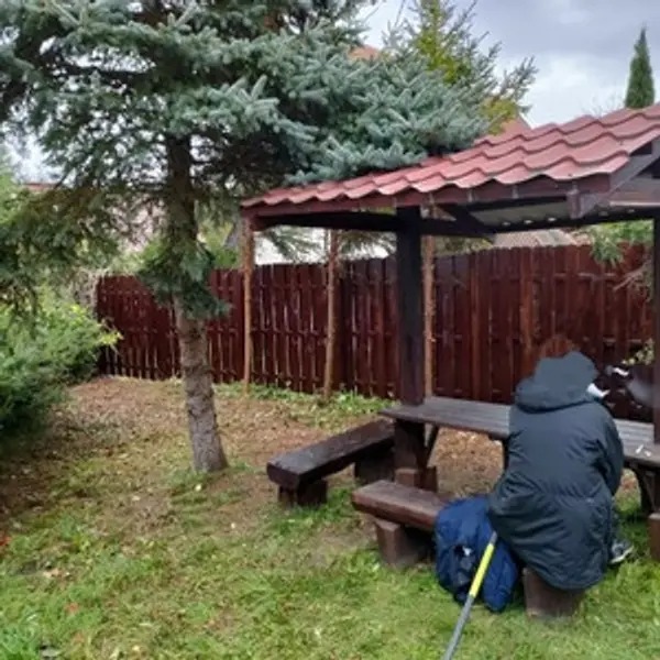 Nowy, ciemnobrązowy płot z desek, widoczny zza drewnianej altany z czerwonym dachem, na tle zielonego trawnika i drzew. Osoba siedzi na ławce w altanie, ubrana w ciemną kurtkę.