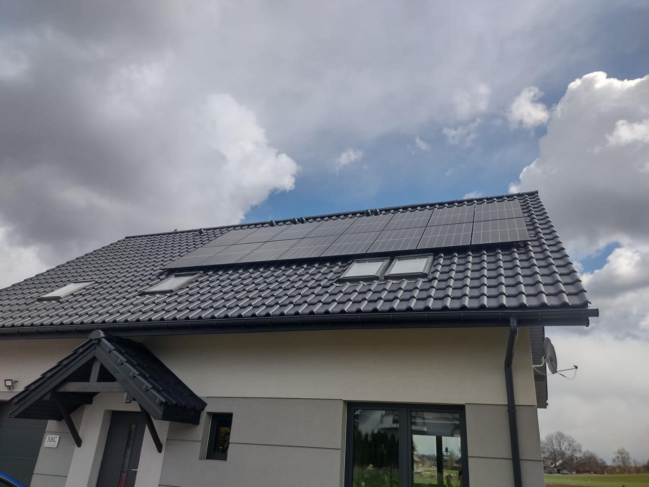 Czarny dach z dachówki ceramicznej z zamontowanymi panelami słonecznymi i dwoma oknami dachowymi, widok z dołu na tle pochmurnego nieba.