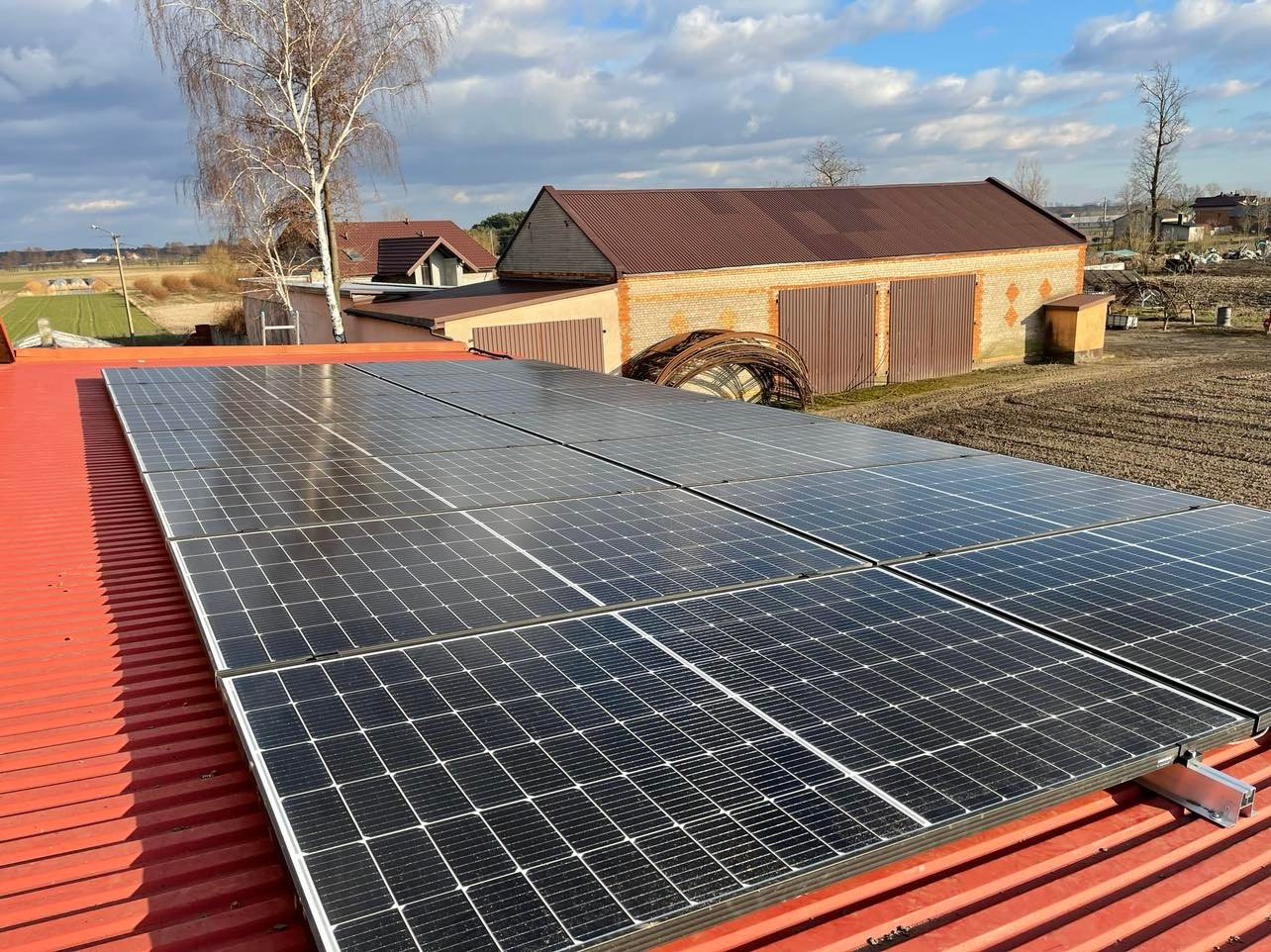 Panele słoneczne zamontowane na czerwonym dachu blaszanym, w tle budynek gospodarczy z cegły i brama garażowa.