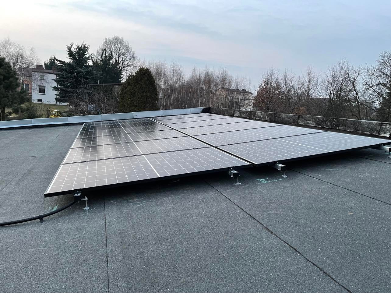 Panele słoneczne zamontowane na płaskim dachu budynku, widoczne elementy mocujące i kable, w tle drzewa i budynek.