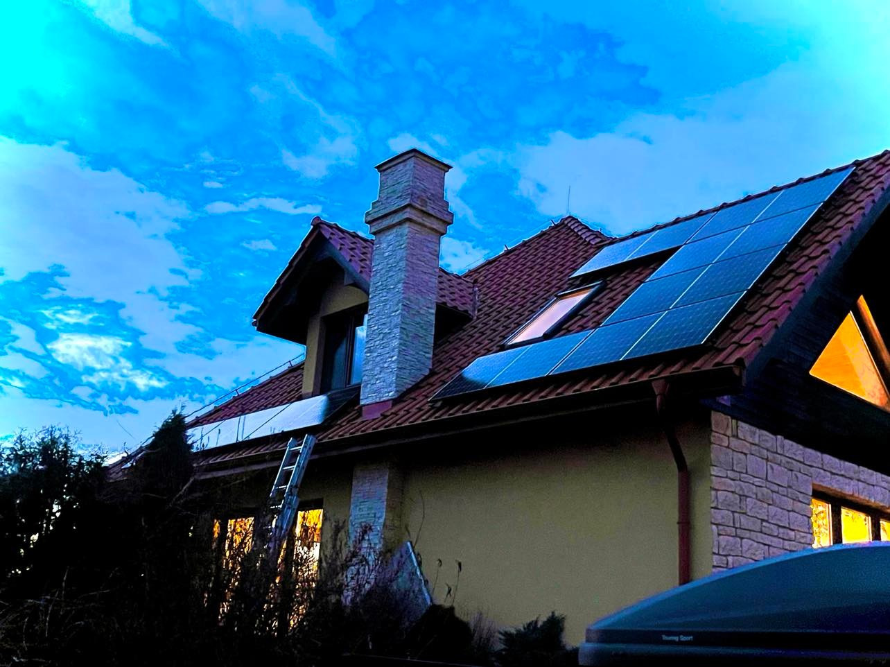 Dom jednorodzinny z zamontowanymi panelami fotowoltaicznymi na dachu z czerwonej dachówki, widoczna drabina oparta o dach podczas instalacji, niebieskie niebo w tle.
