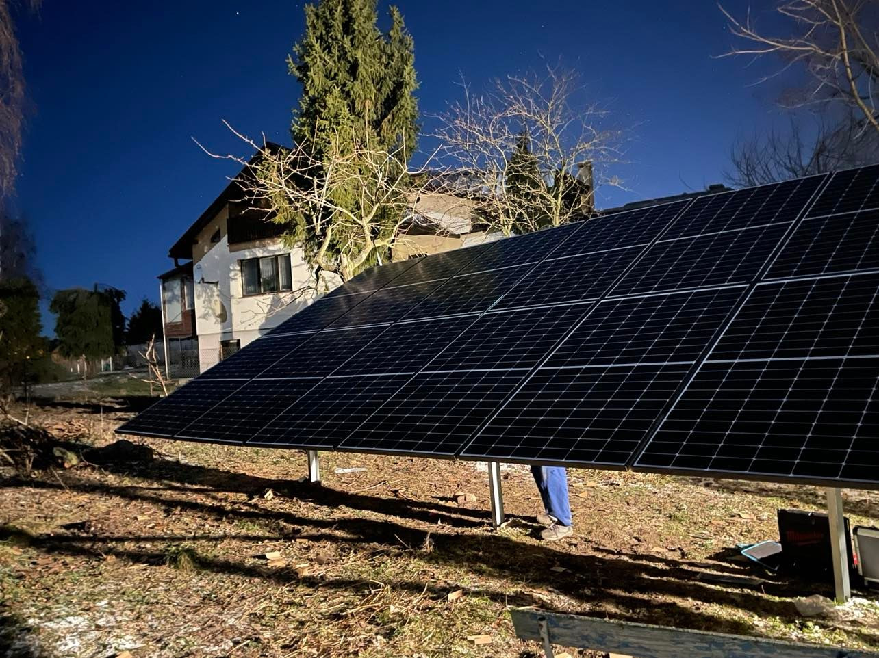 Instalacja paneli fotowoltaicznych na gruncie przy domu jednorodzinnym, widoczne nogi instalatora w niebieskich spodniach, wieczorny krajobraz.