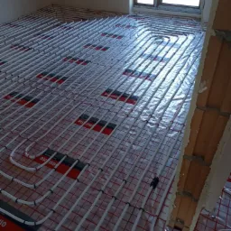 Białe rury ogrzewania podłogowego ułożone na folii z czerwoną kratką, widok z g&oacute;ry, surowy stan, fragment ściany z cegły w tle.