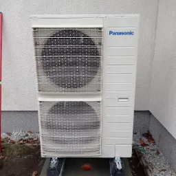 Zewnętrzna jednostka pompy ciepła Panasonic zamontowana na betonowej podstawie z gumowymi podkładkami, widoczne otoczenie budynku z kamieniami i fragmentem elewacji.