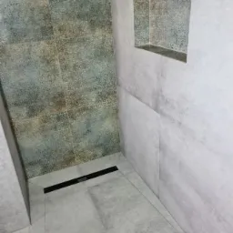 Wykończona kabina prysznicowa z odpływem liniowym i wnęką w ścianie, wyłożona płytkami w odcieniach szarości i beżu z delikatnym wzorem.