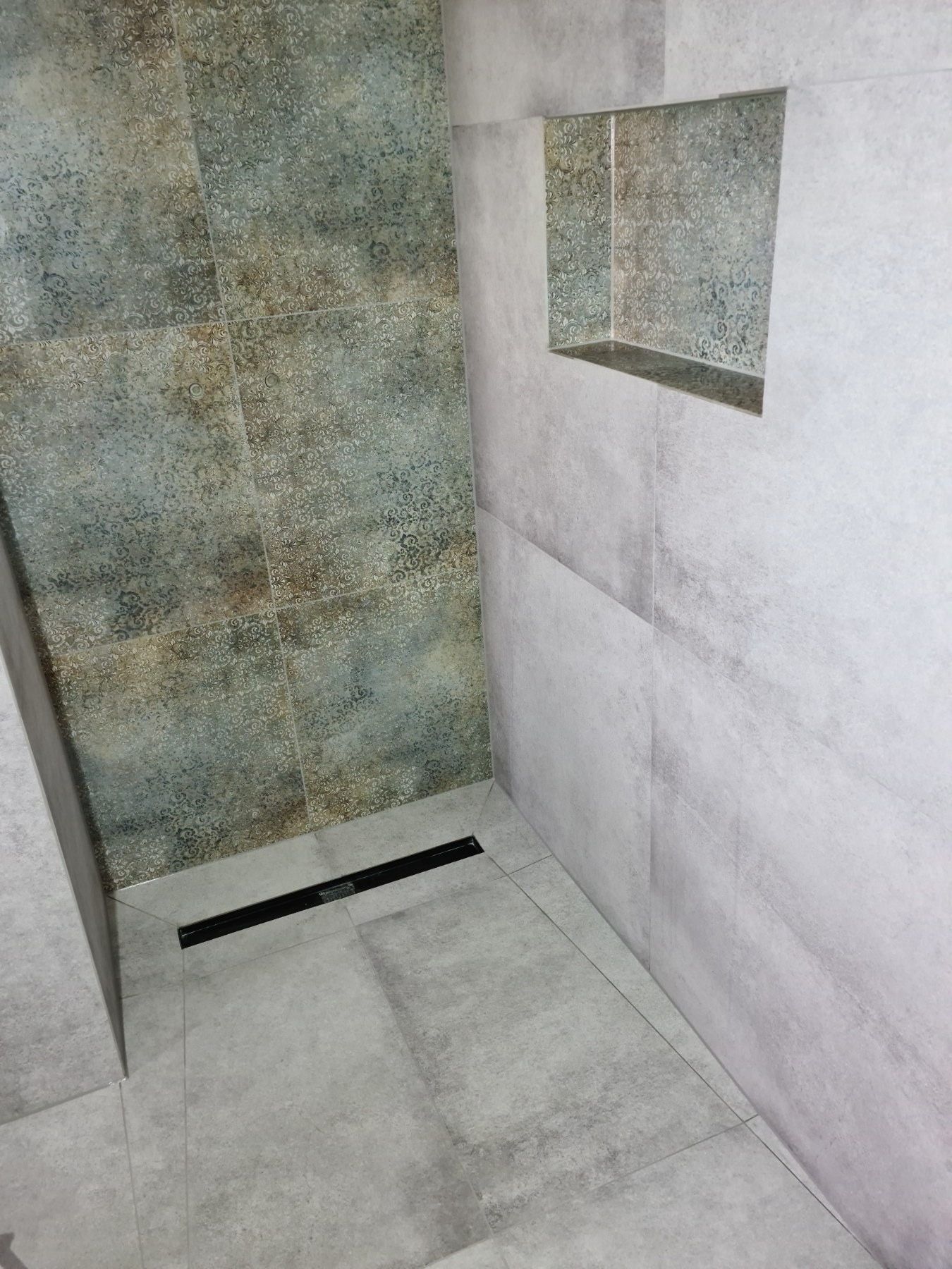 Wykończona kabina prysznicowa z odpływem liniowym i wnęką w ścianie, wyłożona płytkami w odcieniach szarości i beżu z delikatnym wzorem.