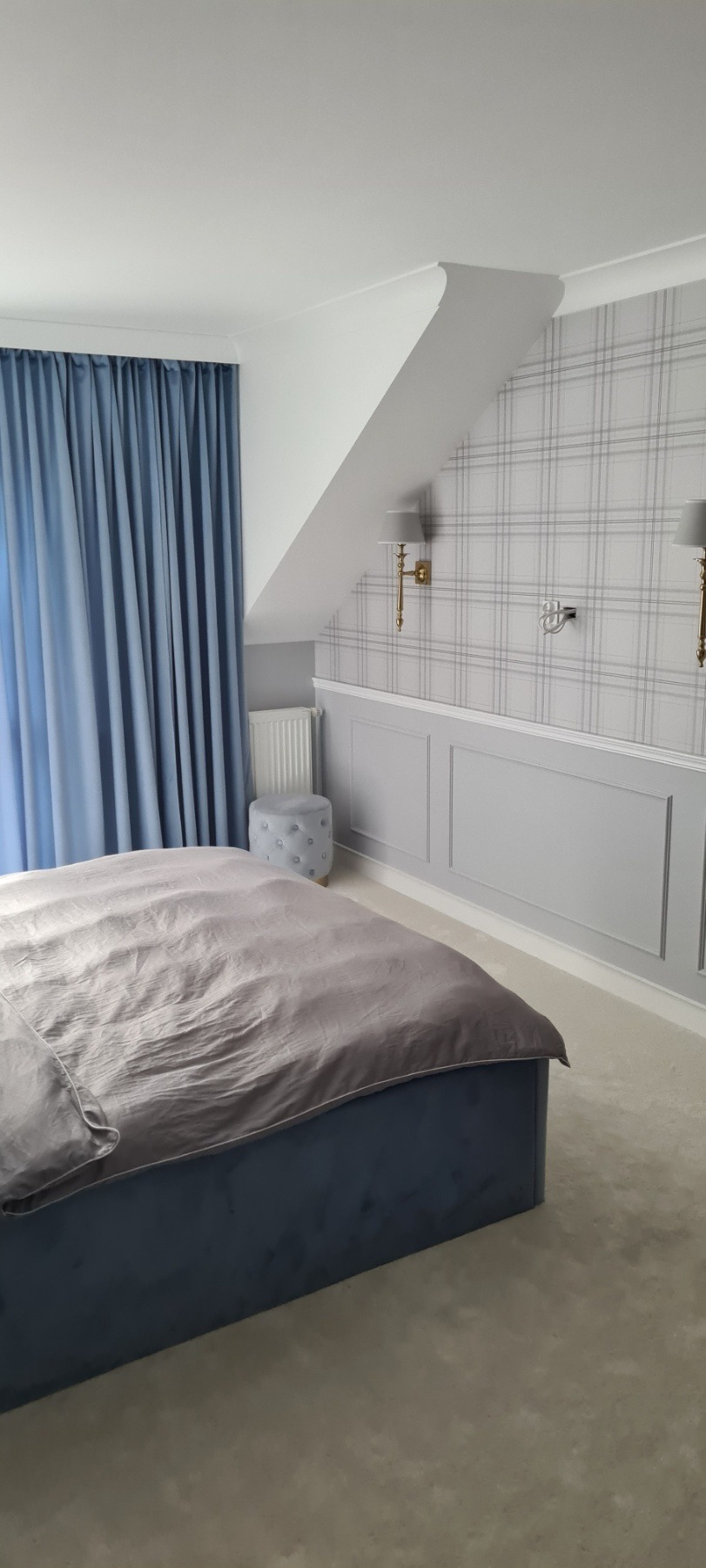 Wykończona sypialnia z łóżkiem z niebieskim zagłówkiem, szarą pościelą, ścianą z szarą tapetą w kratę, szarymi panelami ściennymi, białym sufitem ze skosem i niebieskimi zasłonami.