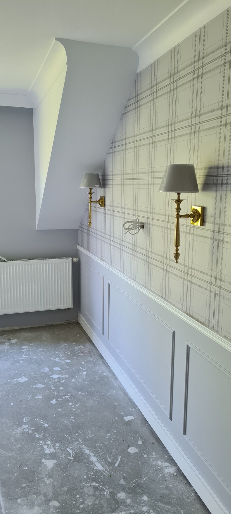 Pokój w trakcie wykańczania: tapeta w szarą kratę, biała boazeria angielska, złote kinkiety z abażurami, ślady farby na podłodze, widoczne przewody elektryczne.