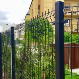 Czarny, metalowy płot panelowy z widocznymi mocowaniami słupków, otaczający posesję z zielenią, na tle dwupiętrowego budynku.