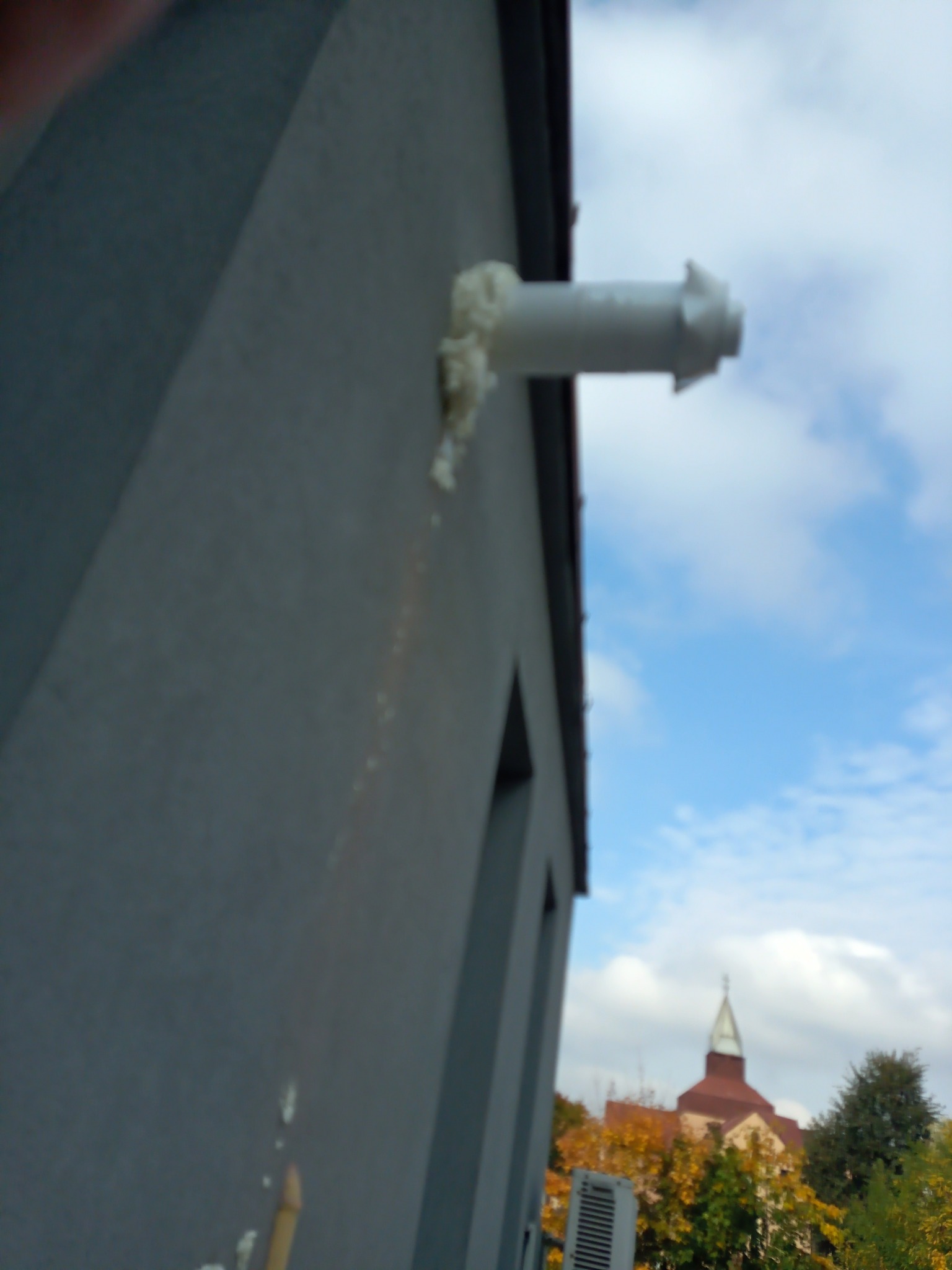 Szary budynek z wystającą rurą wentylacyjną, uszczelnioną pianką montażową, widok na kościół i drzewa w tle, pochmurne niebo.