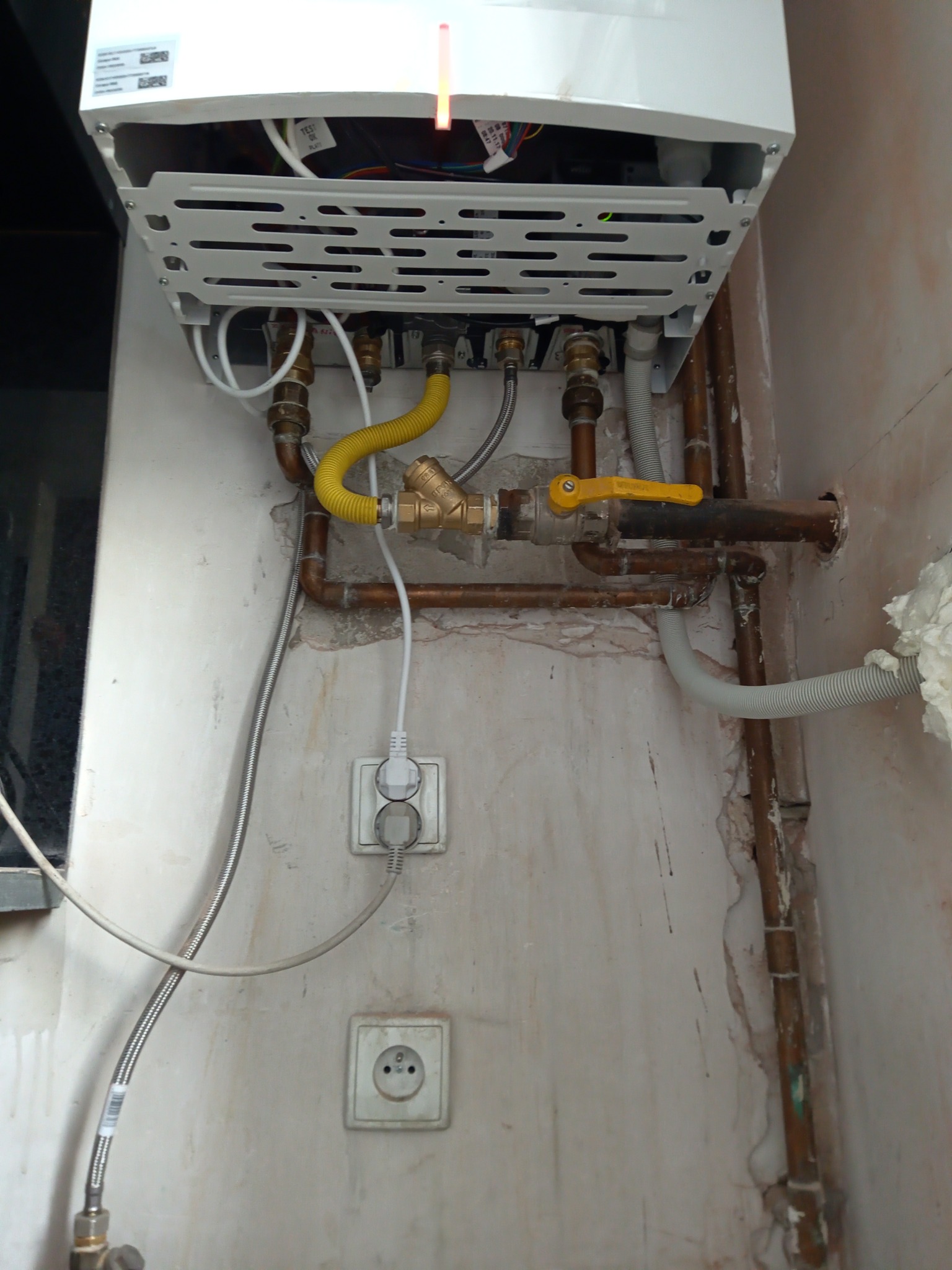 Biały piec gazowy zamontowany na ścianie z widocznymi rurami miedzianymi, żółtym zaworem i elastycznym przewodem gazowym, gniazdka elektryczne w pobliżu, ściana w stanie surowym.