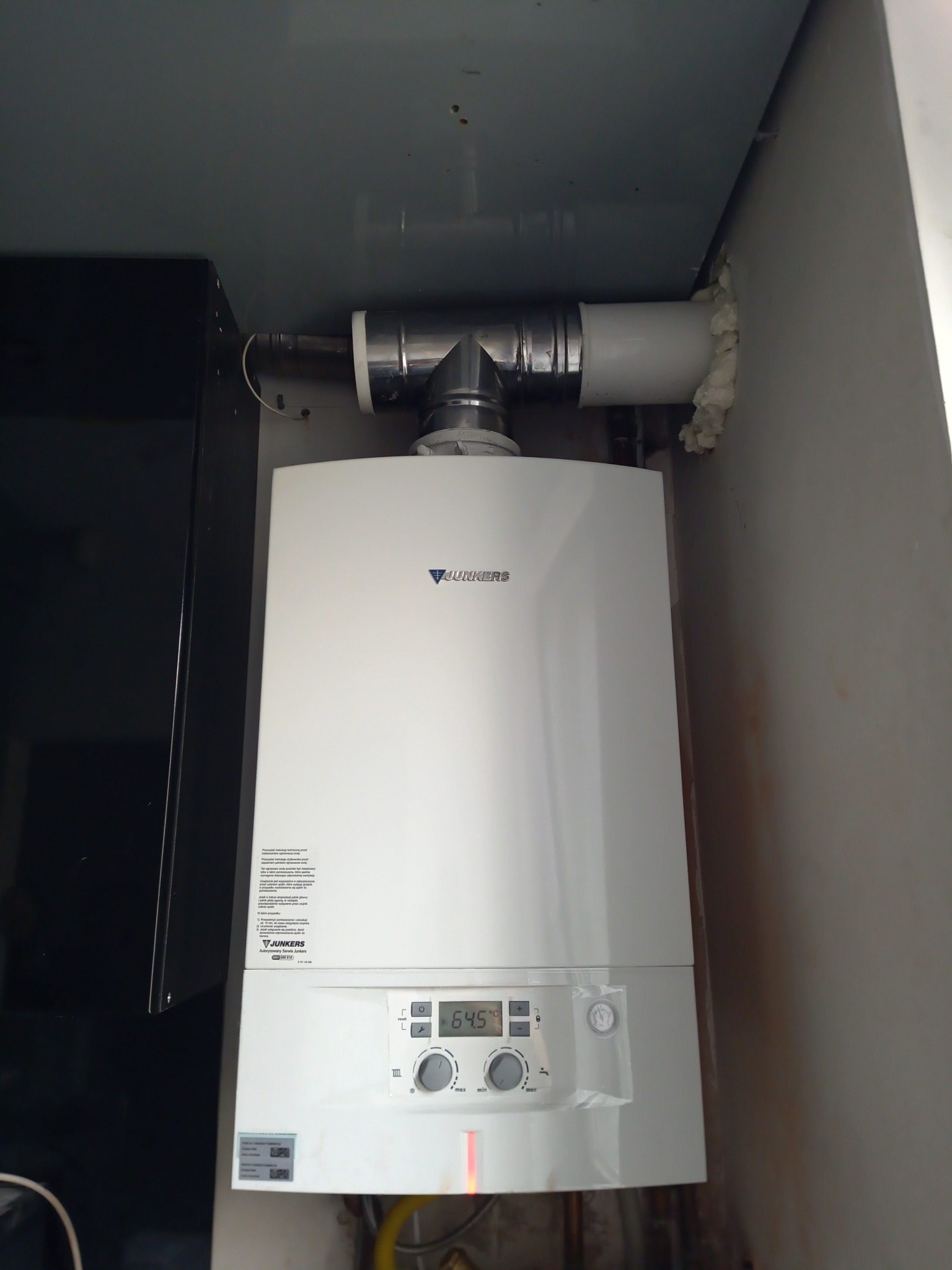Biały piec gazowy Junkers z podłączonymi rurami spalinowymi, zamontowany w wąskiej przestrzeni między ścianą a czarną szafką, uszczelniony pianką montażową przy ścianie.