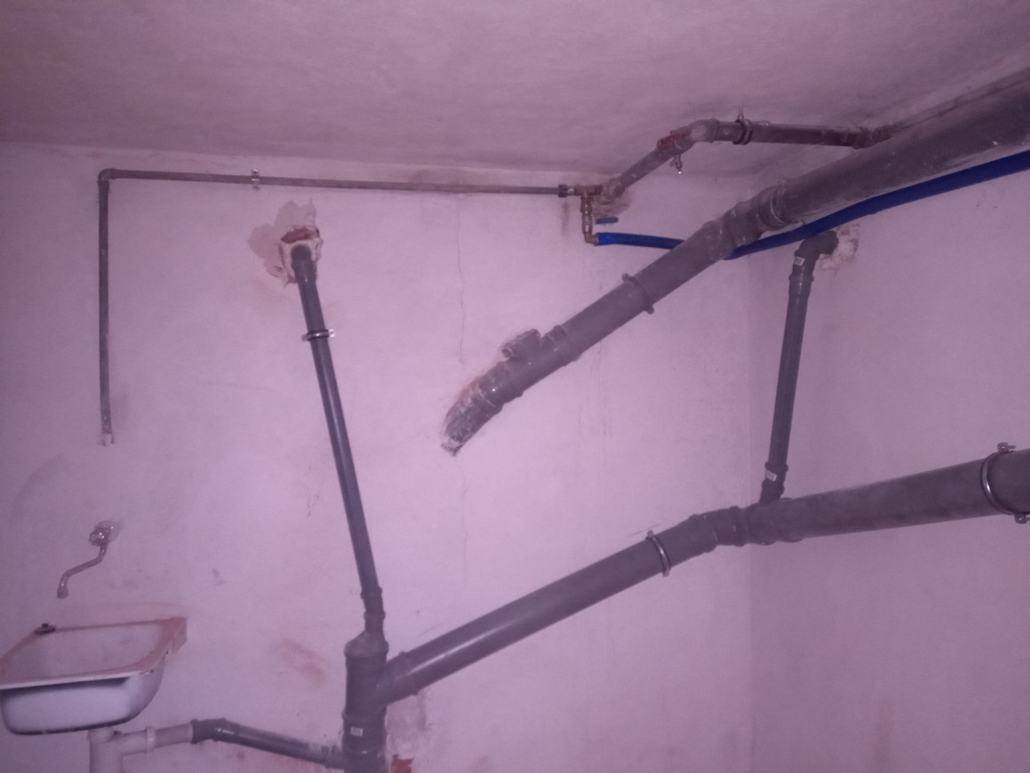 Szare rury kanalizacyjne z metalowymi obejmami, niebieska rurka i zawory w surowym pomieszczeniu z umywalką, widoczne ślady po kuciu w ścianach.