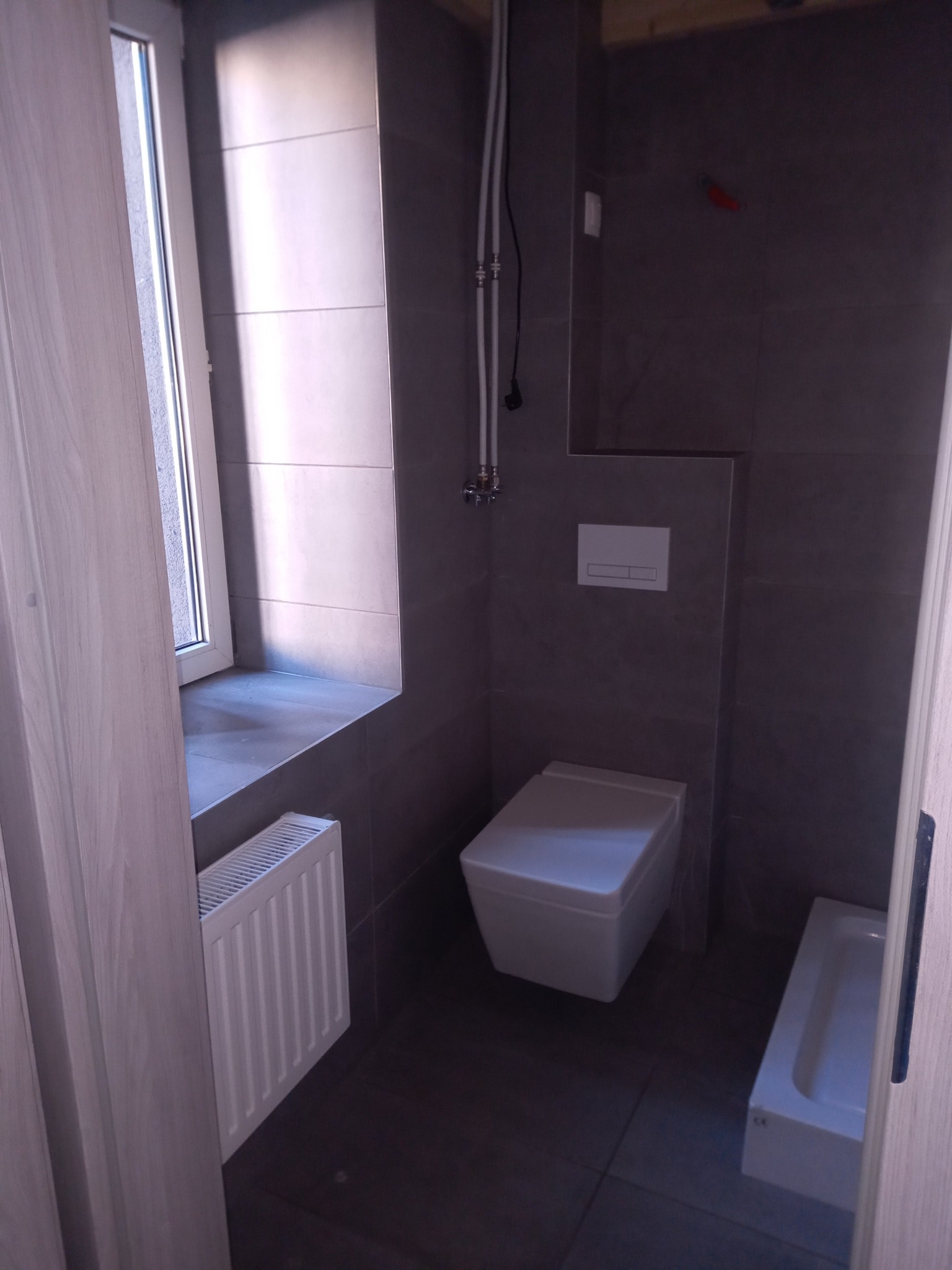 Szara łazienka z podwieszaną toaletą, grzejnikiem pod oknem i fragmentem wanny, widok zza częściowo otwartych drzwi.
