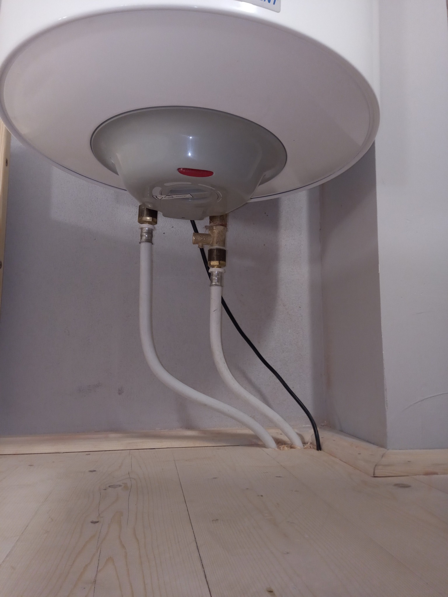 Podłączenie bojlera: biały bojler z widocznymi rurami doprowadzającymi wodę i kablem zasilającym, zamontowany przy szarej ścianie i drewnianej podłodze.