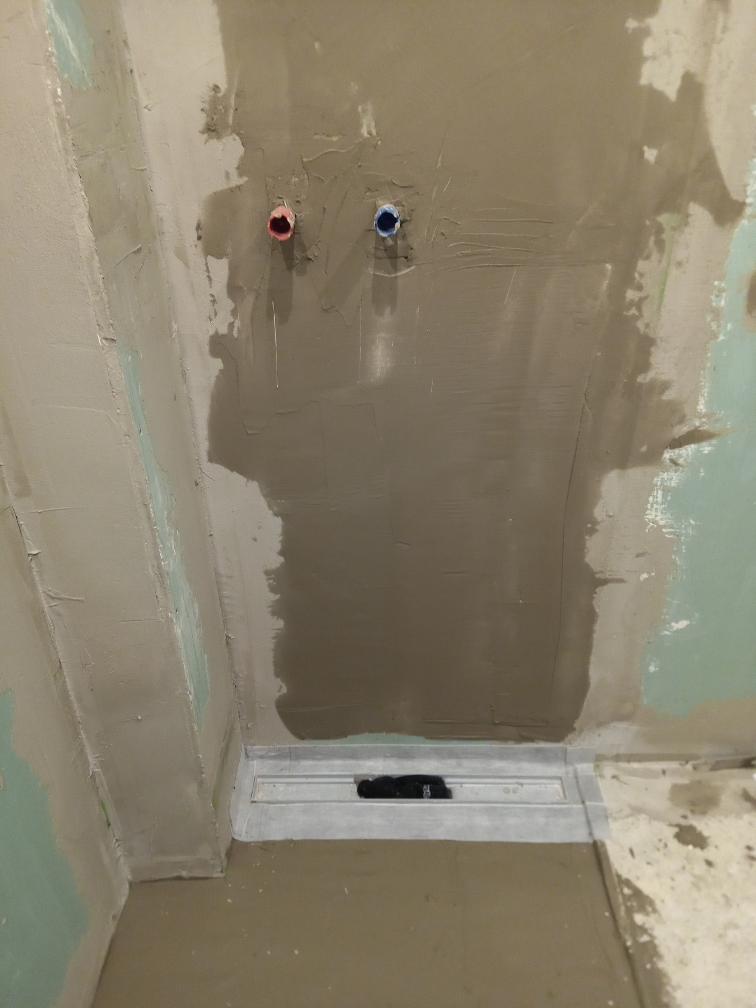 Nierówna ściana w trakcie remontu łazienki z widocznymi rurami wodnymi (czerwona i niebieska) i odpływem liniowym w posadzce, pokryta świeżym tynkiem.