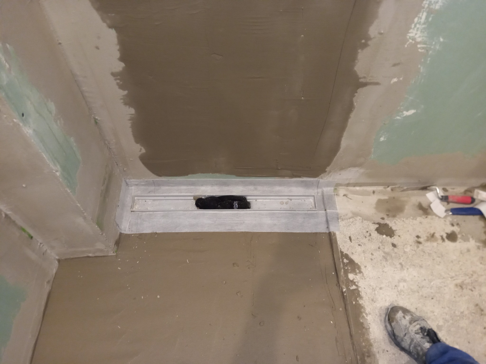 Nierównomiernie nałożona wylewka samopoziomująca w łazience, widoczny odpływ liniowy i narzędzia malarskie w tle, stopa w roboczym obuwiu w prawym dolnym rogu.