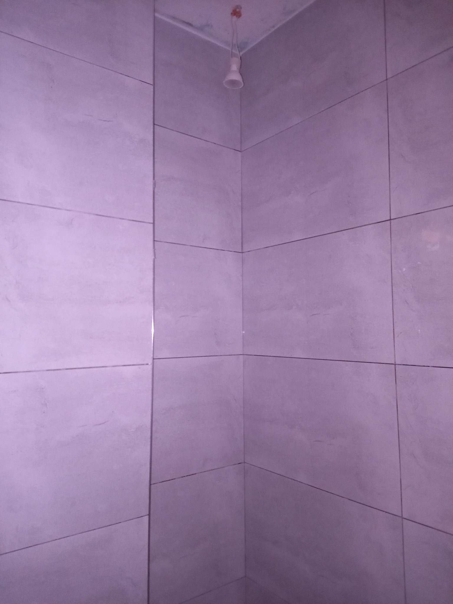 Wykończony róg łazienki z ułożonymi, szarymi płytkami ceramicznymi o matowym wykończeniu; widoczna surowa żarówka na kablu.