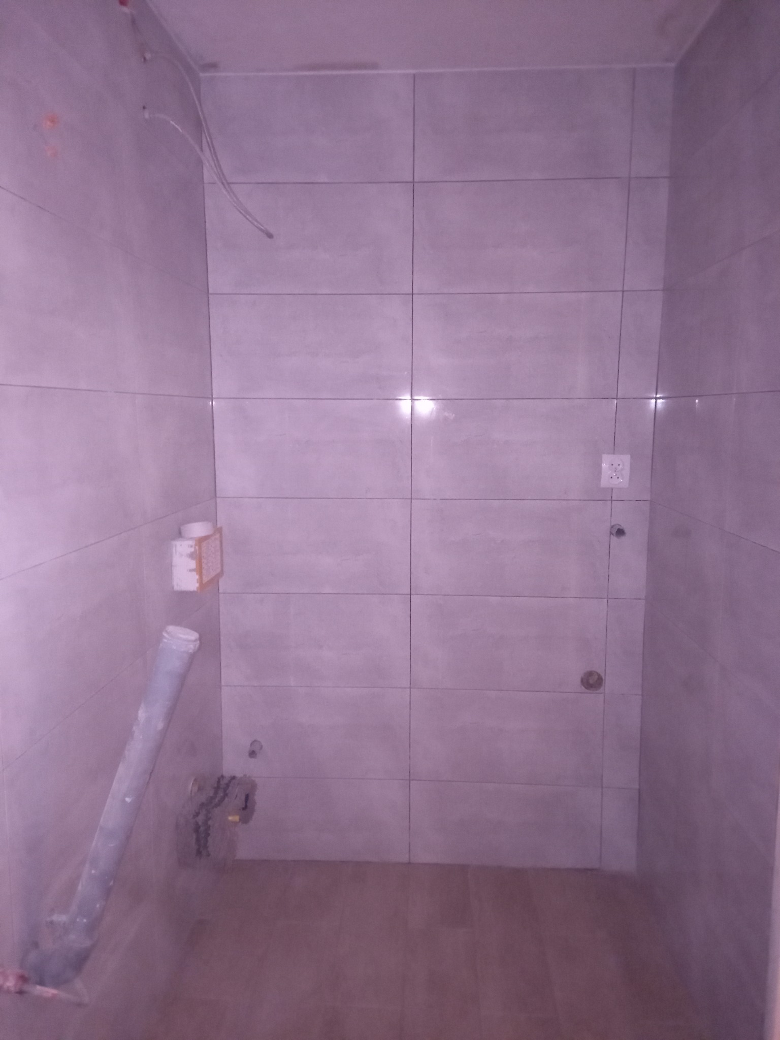 Wnętrze łazienki w trakcie remontu, widoczne szare płytki na ścianach, wystające kable, rura kanalizacyjna w rogu oraz gniazdko elektryczne.