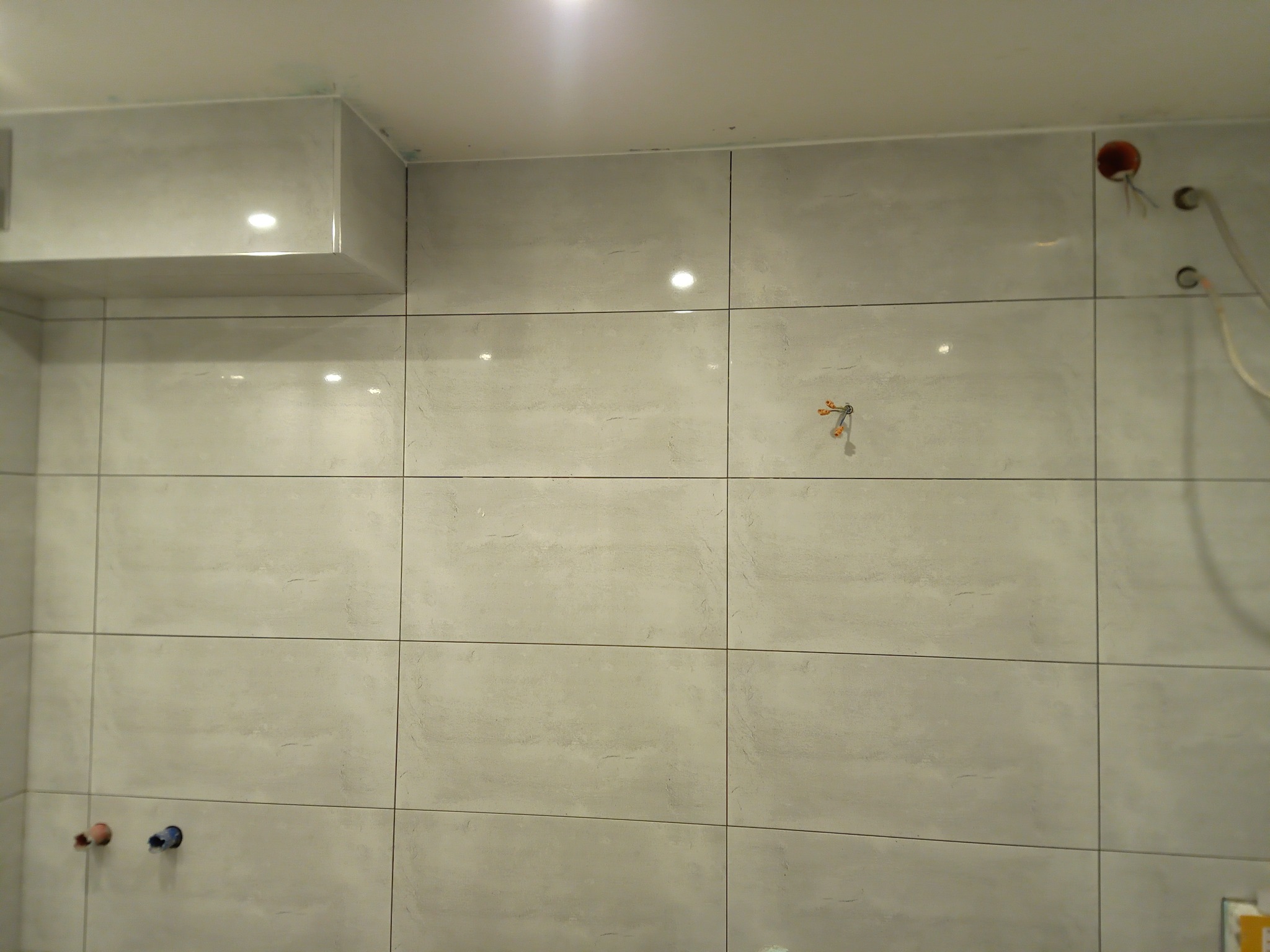 Ściana łazienki wyłożona jasnymi, matowymi płytkami ceramicznymi w trakcie prac wykończeniowych, widoczne wystające przewody elektryczne i rury instalacyjne.