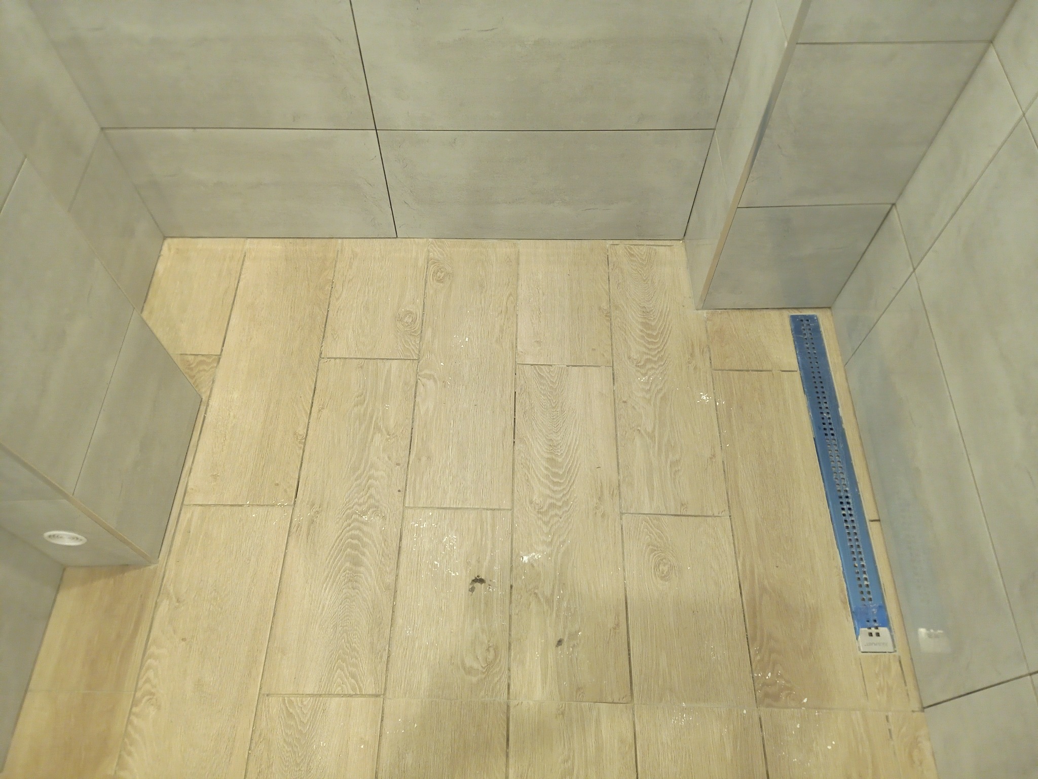 Wykończona kabina prysznicowa z odpływem liniowym, wyłożona płytkami imitującymi drewno na podłodze i szarymi płytkami na ścianach.