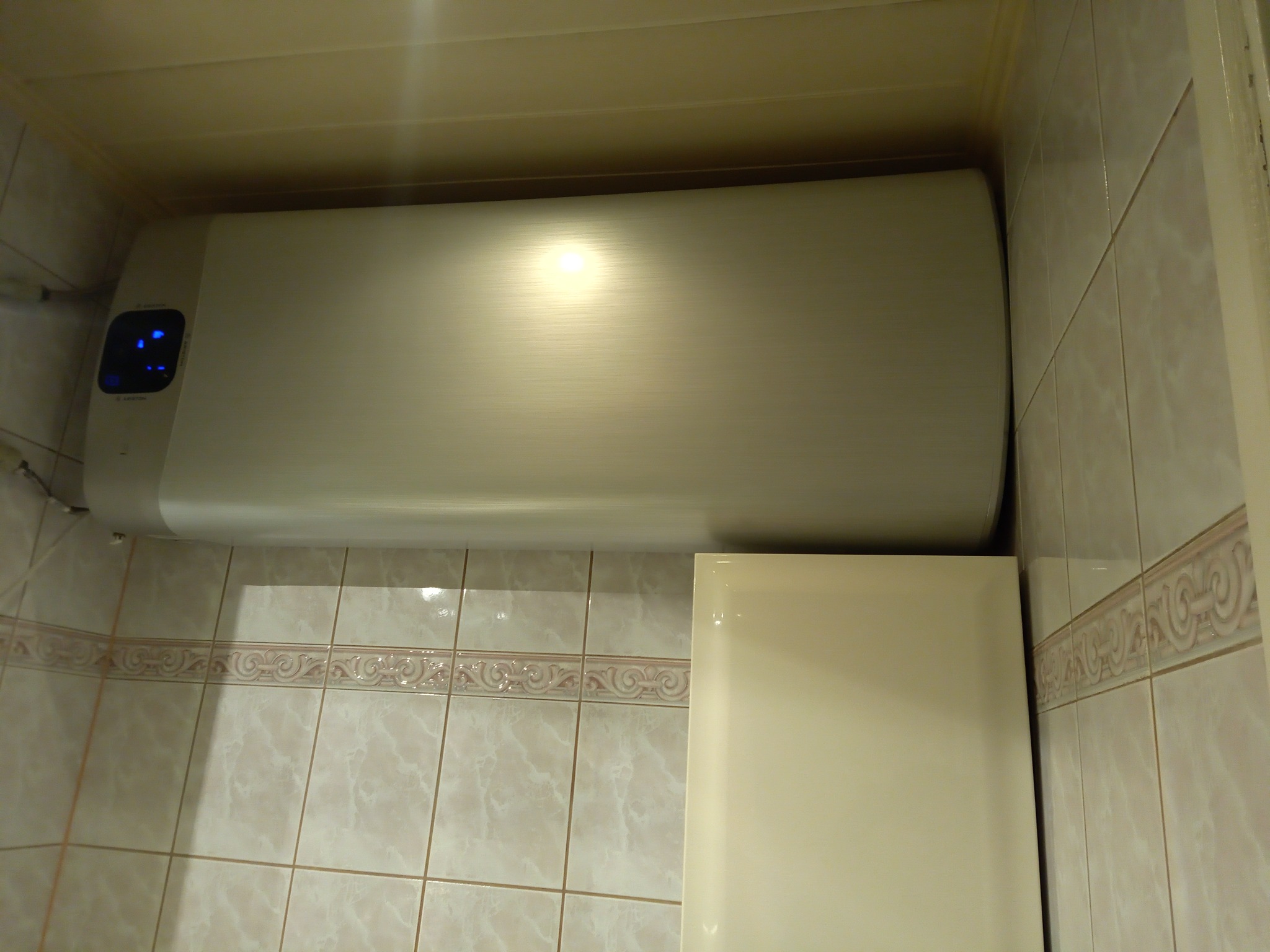 Poziomy, elektryczny podgrzewacz wody zamontowany w łazience z beżowymi, marmurkowymi płytkami i ozdobnym pasem na ścianie.