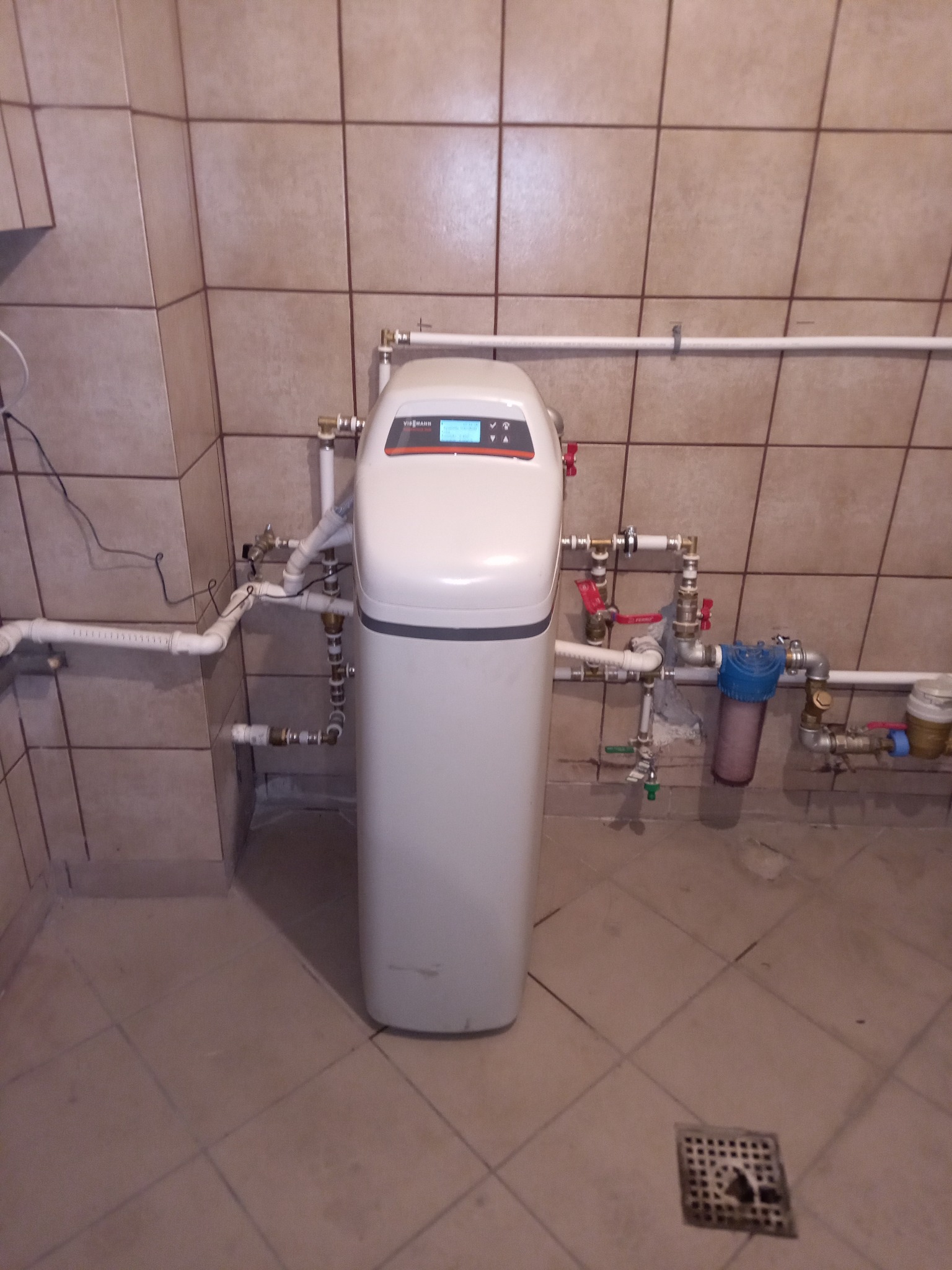 Biały zmiękczacz wody Viessmann zamontowany w pomieszczeniu z beżowymi płytkami, widoczne rury i zawory, filtr wody z niebieską obudową oraz kratka ściekowa w podłodze.
