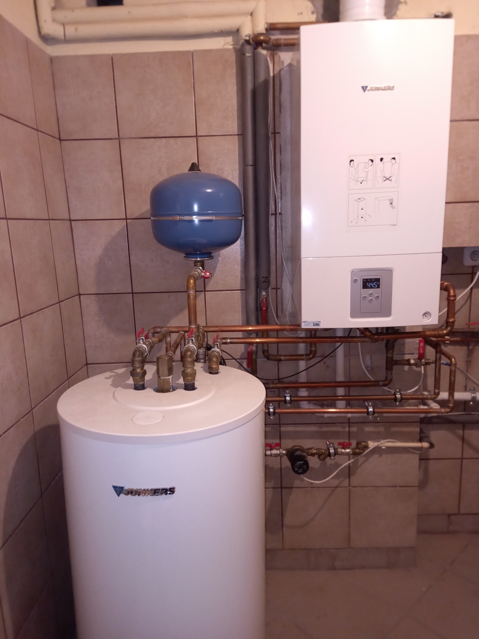 Bojler i piec gazowy Junkers z instalacją hydrauliczną na tle płytek ceramicznych w kotłowni.