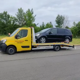Żółta laweta z czarnym samochodem osobowym na platformie, zabezpieczonym pasami transportowymi, widoczny kierowca lawety
