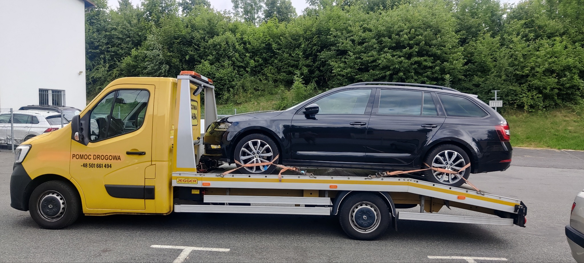 Czarny samochód kombi zabezpieczony pasami transportowymi na żółtej lawecie z napisem 'Pomoc Drogowa' i numerem telefonu.