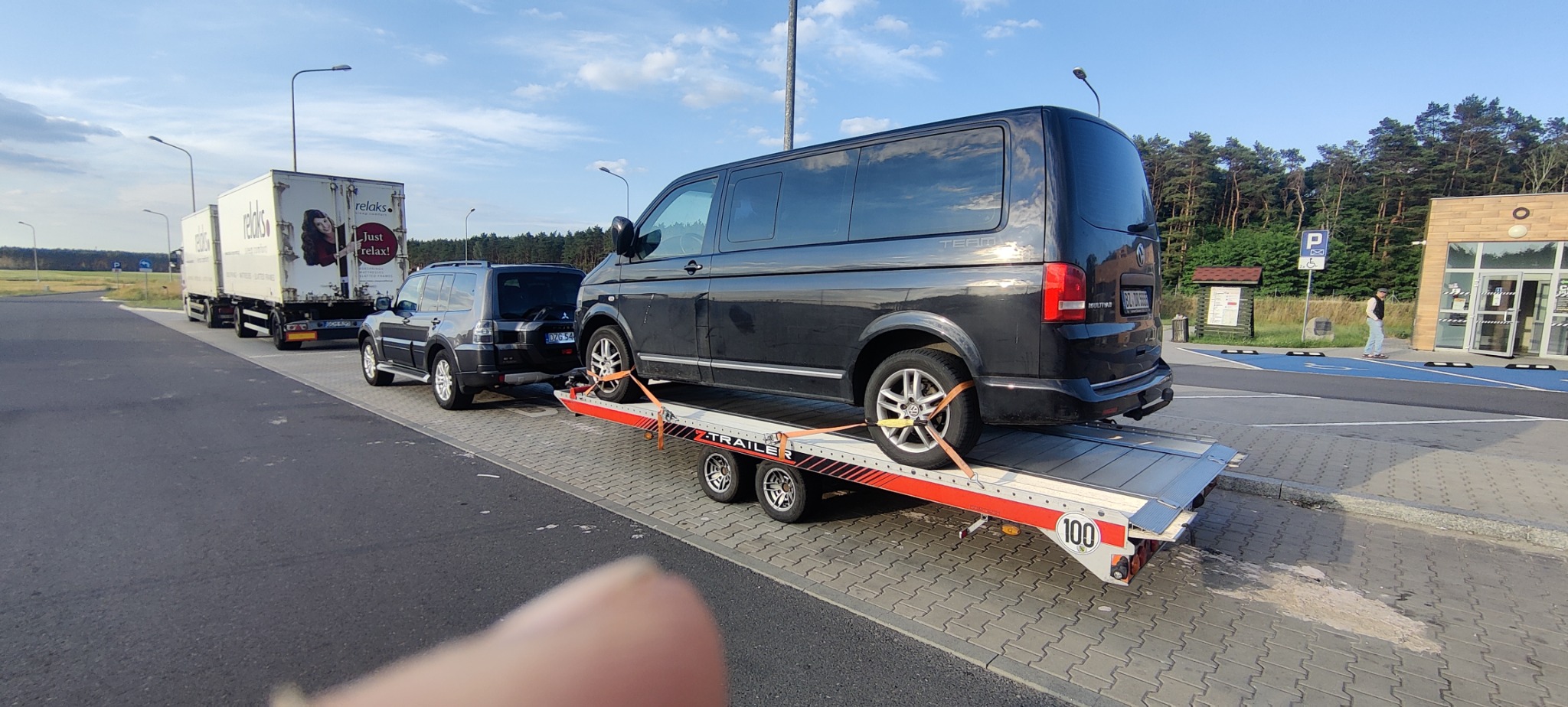 Czarny van Volkswagen T5 zabezpieczony pasami na platformie lawety, za nią holujący ciemny SUV Mitsubishi Pajero, w tle parking z ciężarówką i budynkiem.