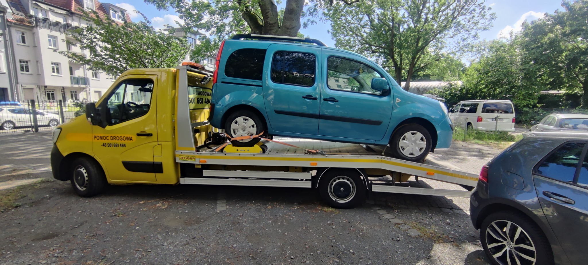 Żółta laweta z niebieskim samochodem osobowym na platformie, zabezpieczonym pasami transportowymi. W tle budynki mieszkalne i zieleń drzew.