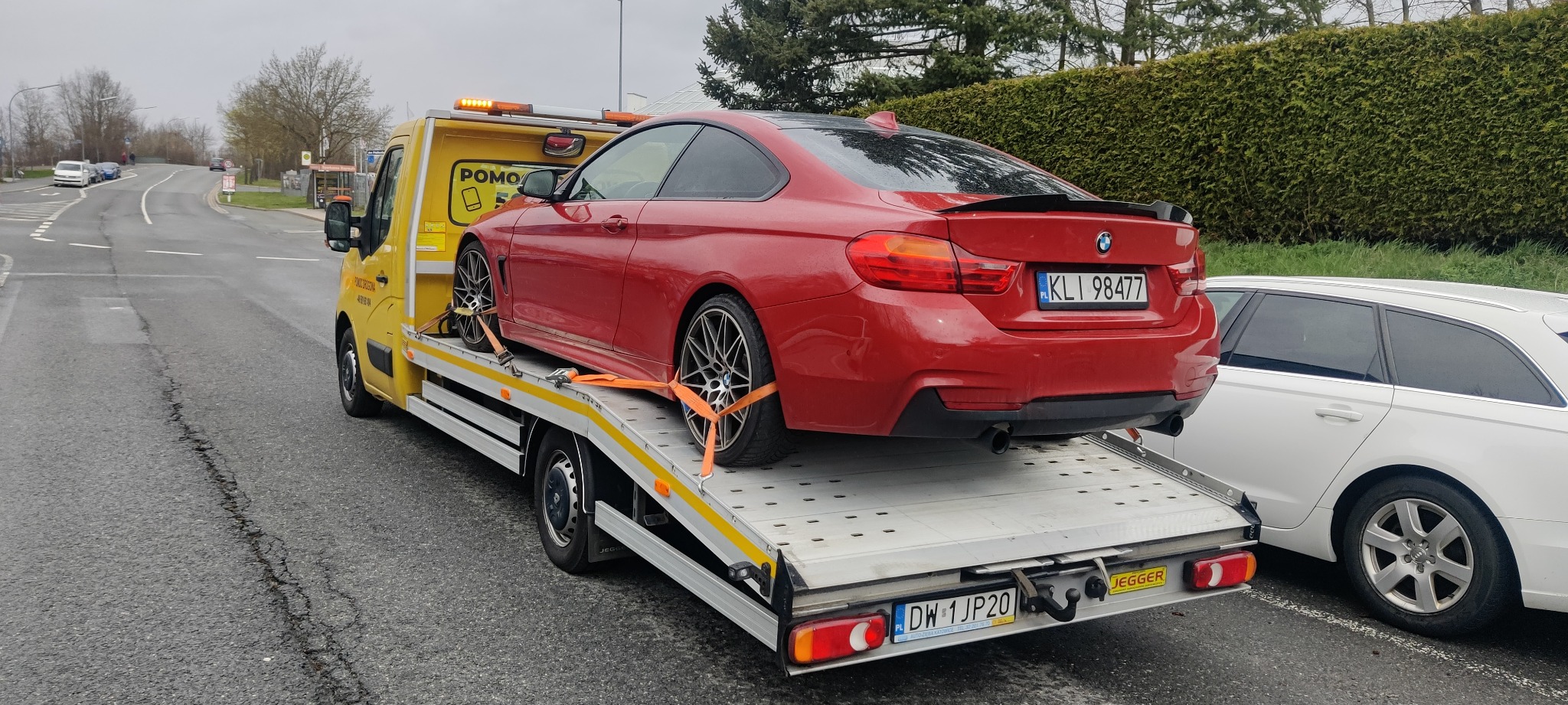 Czerwone BMW serii 4 zabezpieczone pomarańczowymi pasami na platformie żółtej ciężarówki pomocy drogowej z włączonymi światłami ostrzegawczymi, widoczne tablice rejestracyjne.