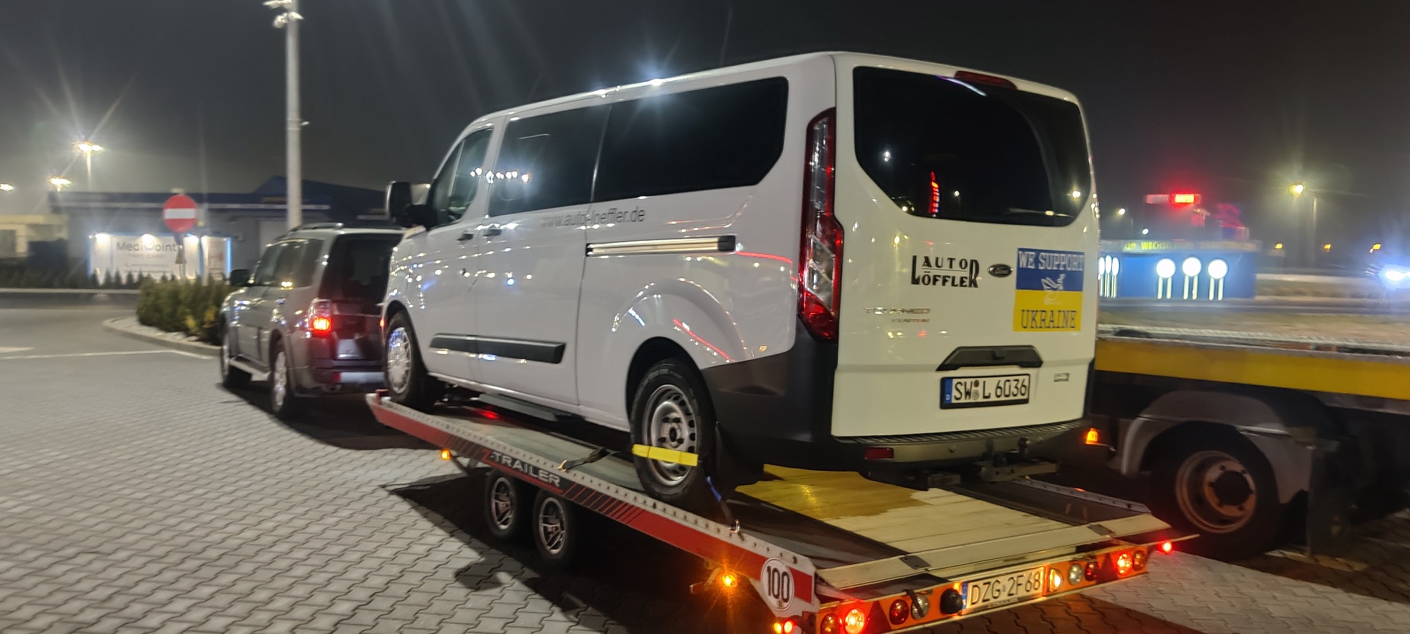 Biały bus Ford Transit zabezpieczony pasami transportowymi na platformie lawety, widoczny z tyłu, z naklejką 'We support Ukraine', tablice rejestracyjne SW L 6036.