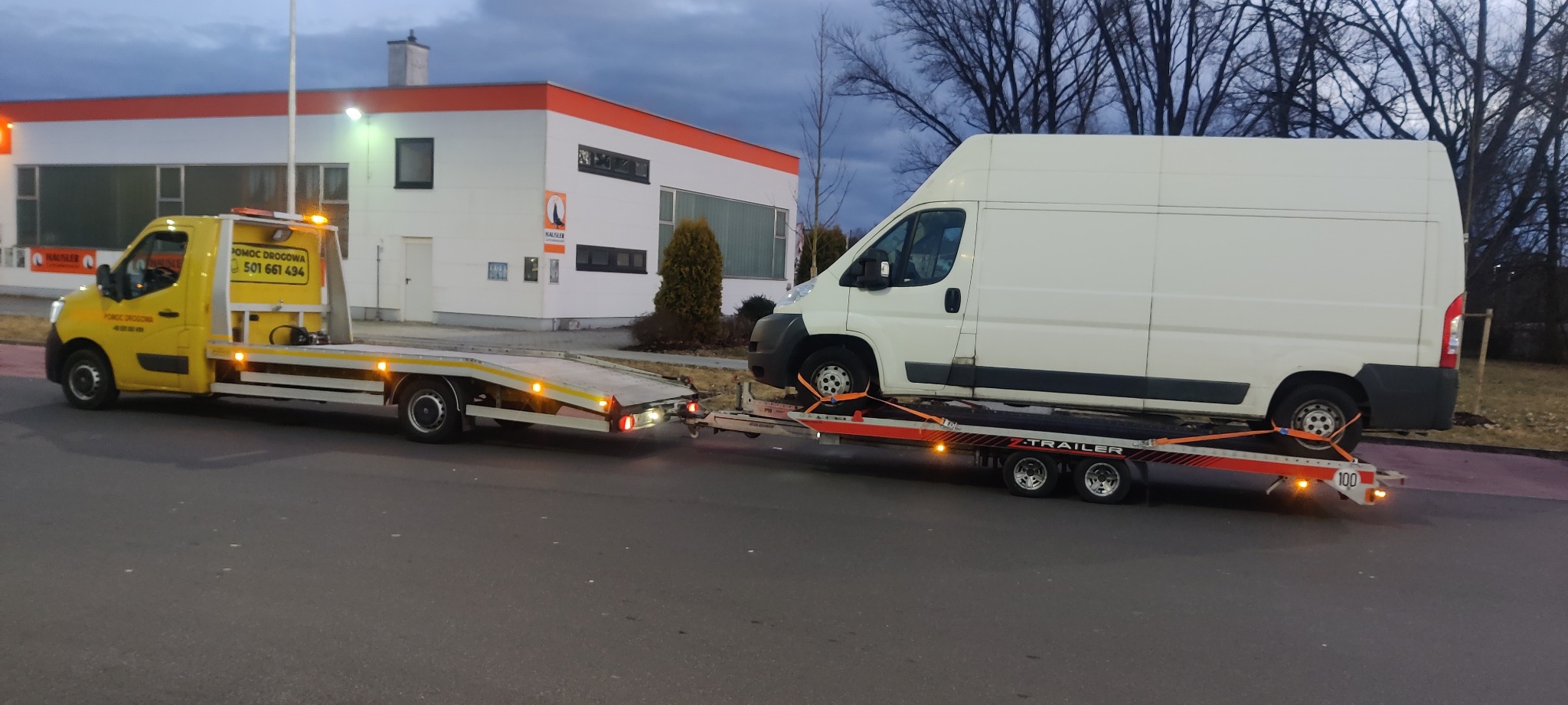 Żółta laweta z pomarańczowymi światłami przewozi biały bus na platformie z czerwonymi pasami mocującymi, na tle budynku z pomarańczowym dachem.