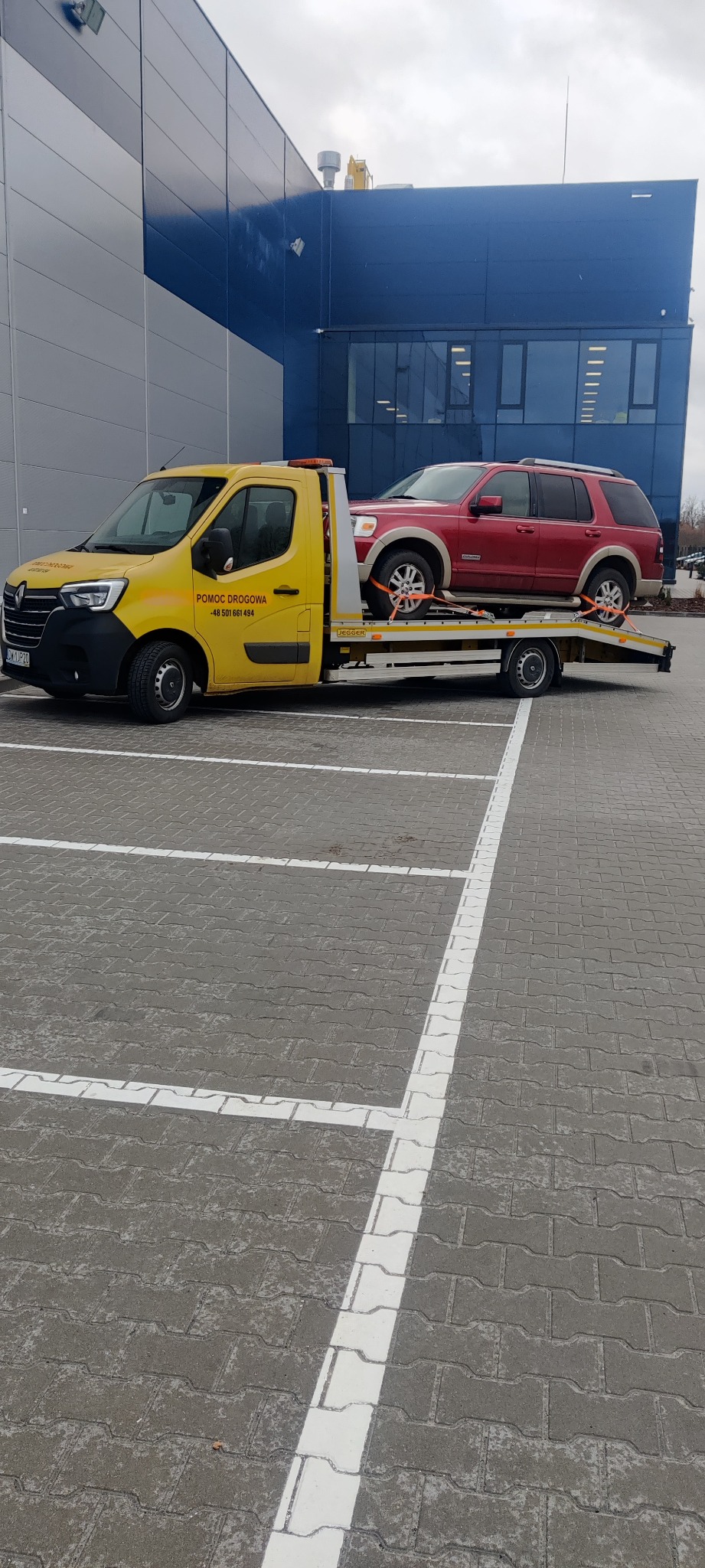 Żółta laweta z czerwonym SUV-em na platformie, zabezpieczonym pomarańczowymi pasami transportowymi, stoi na parkingu w Zgorzelcu, na tle nowoczesnego budynku.